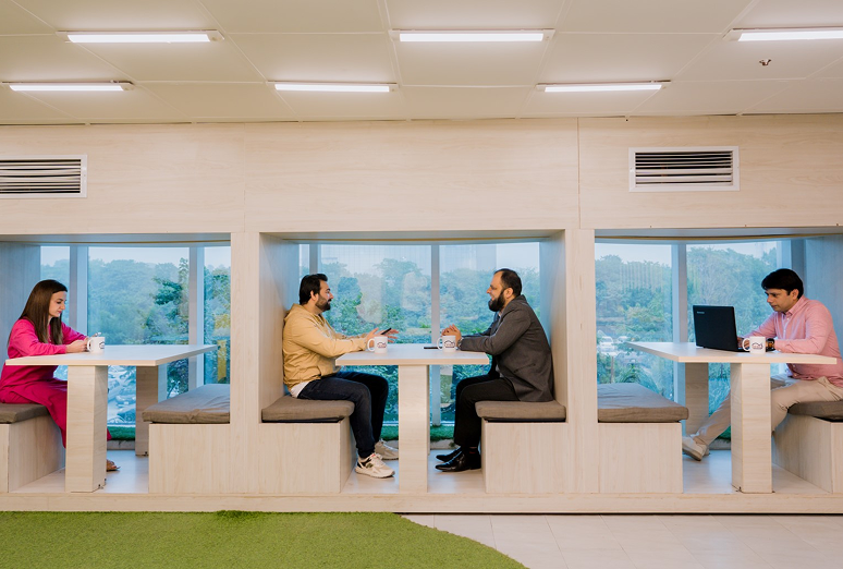 Four people sitting at separate tables in a modern office or cafe with large windows, greenery outside, and a green carpeted area in the foreground.