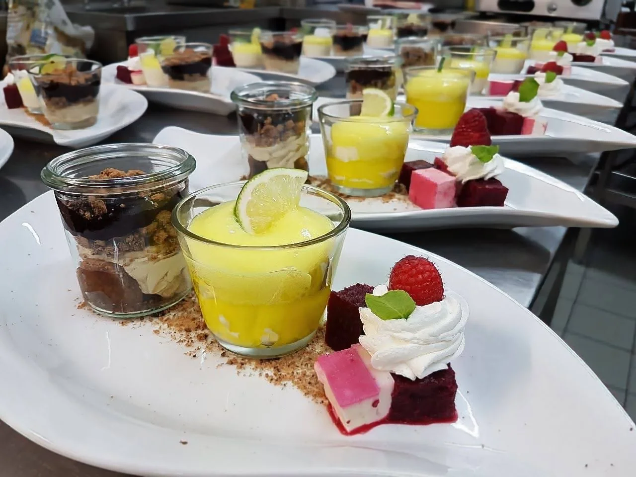 Verschiedene kleine Desserts auf weißen Tellern, darunter Kuchen, Fruchtgelee, Schlagsahne mit Himbeere, Minzblatt, und Schokoladenmousse