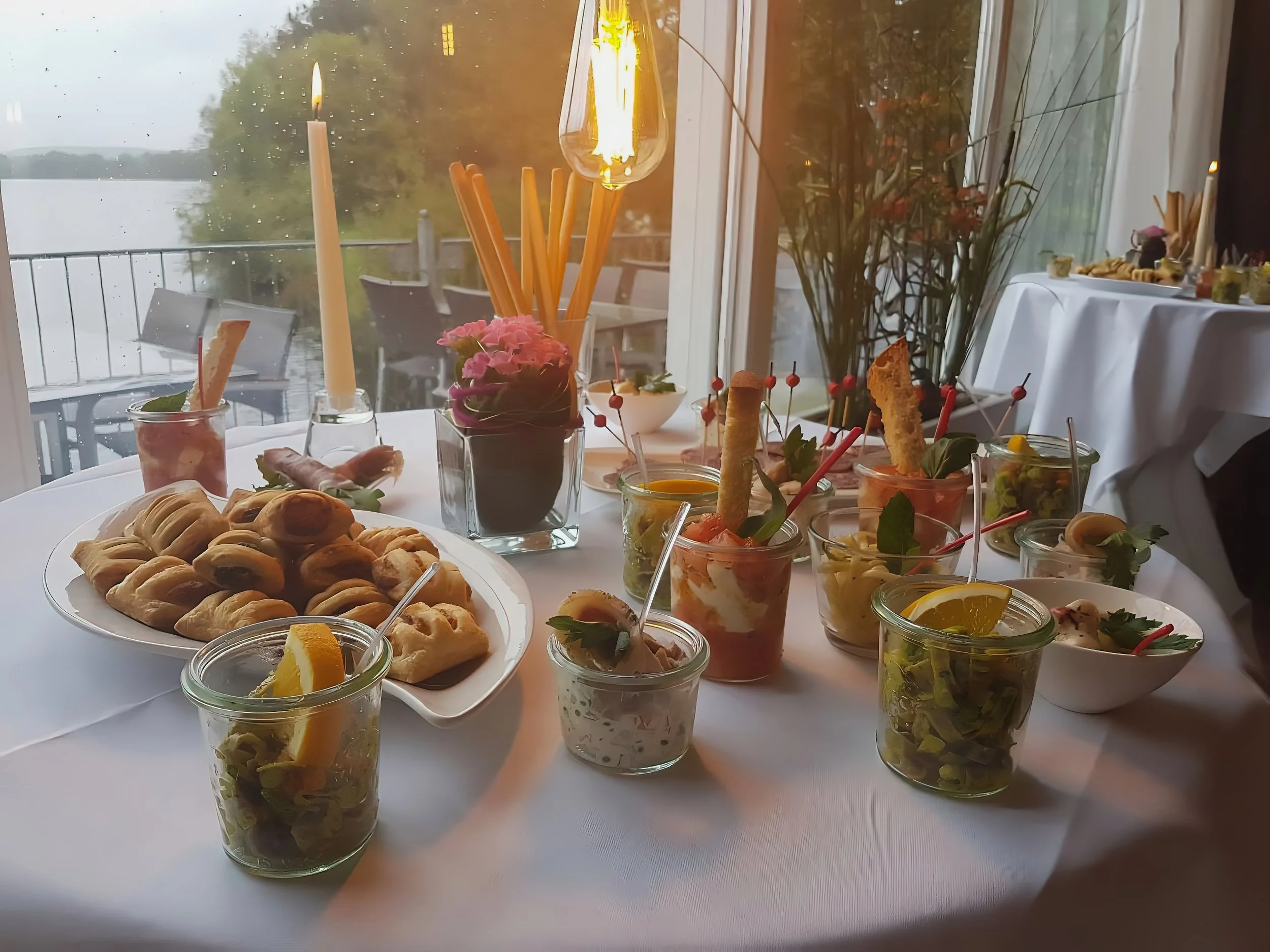 Ein festlich gedeckter Stehtisch mit Fingerfood. Vorspeisen, Salaten, Oliven, Grissini, Kerzen und Dekorationen vor einem Fenster mit Blick auf einen See