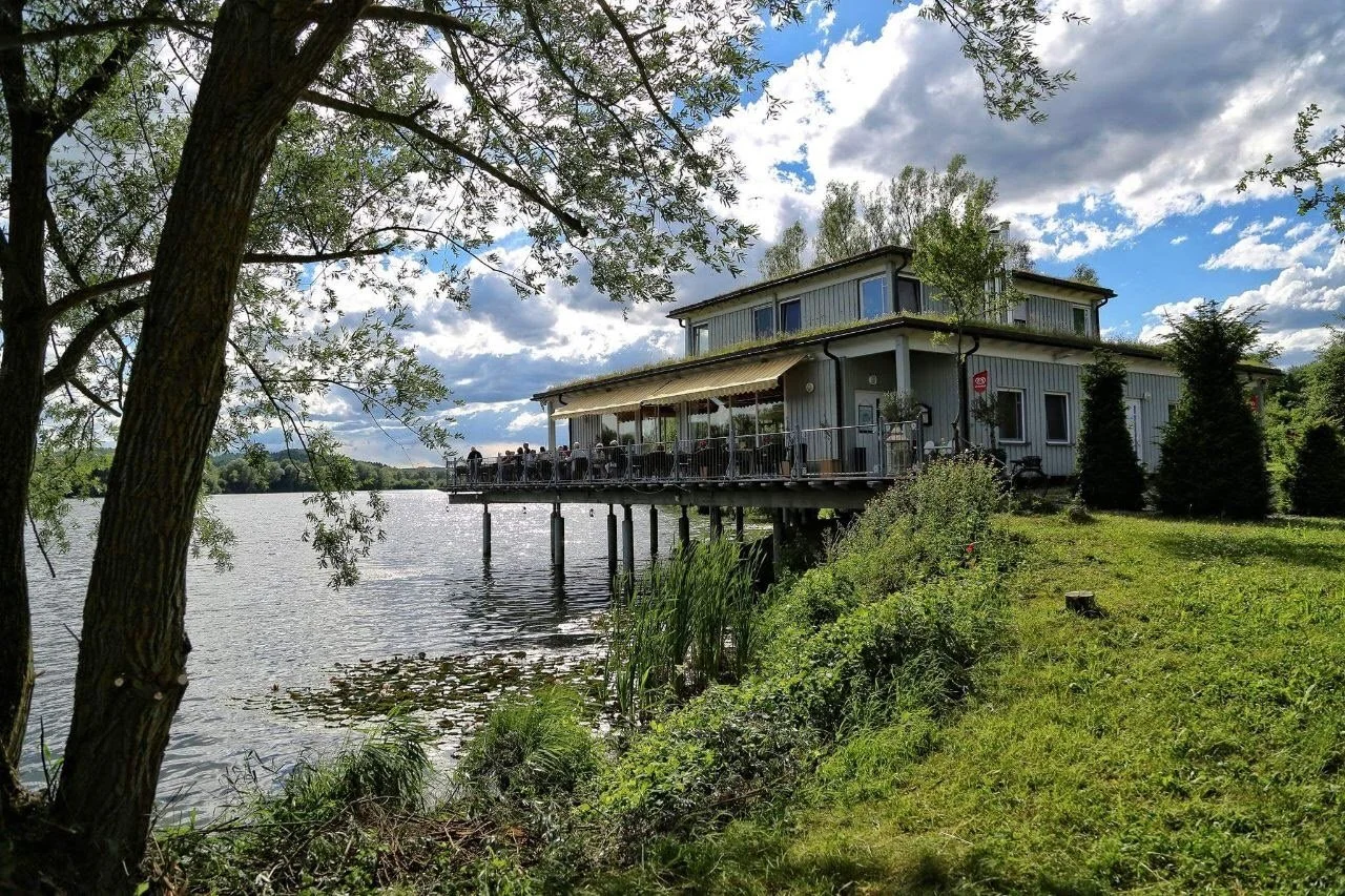Ein Haus auf Stelzen am Ufer eines Sees, umgeben von Bäumen und Gras, mit einem großen Balkon, auf dem Menschen sitzen. Wolkiger Himmel mit Sonnenlicht.