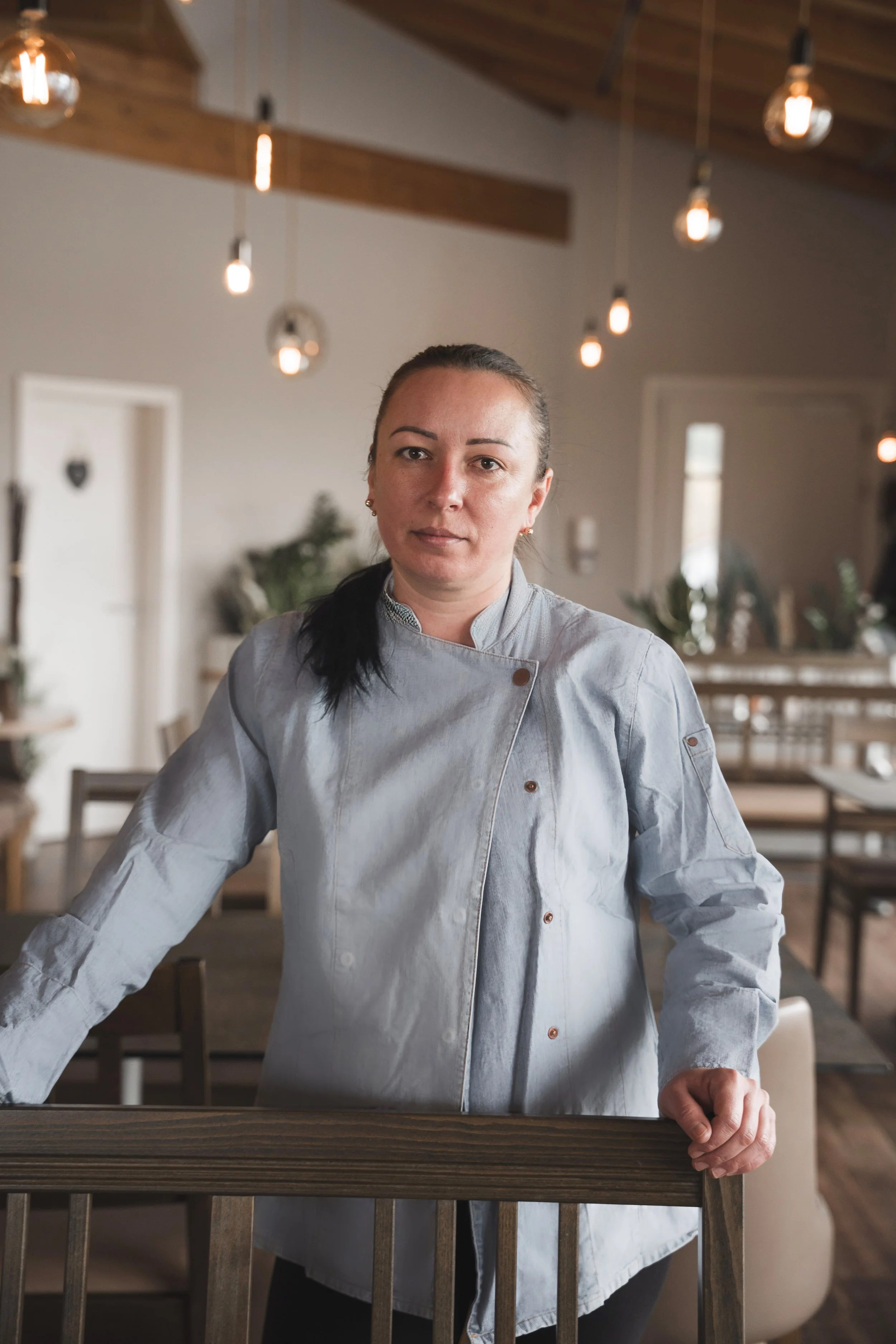 Eine Frau in einer Kochjacke steht in einem modern eingerichteten Restaurant. Sie schaut ernst in die Kamera, im Hintergrund hängen dekorative Glühbirnen an der Decke.