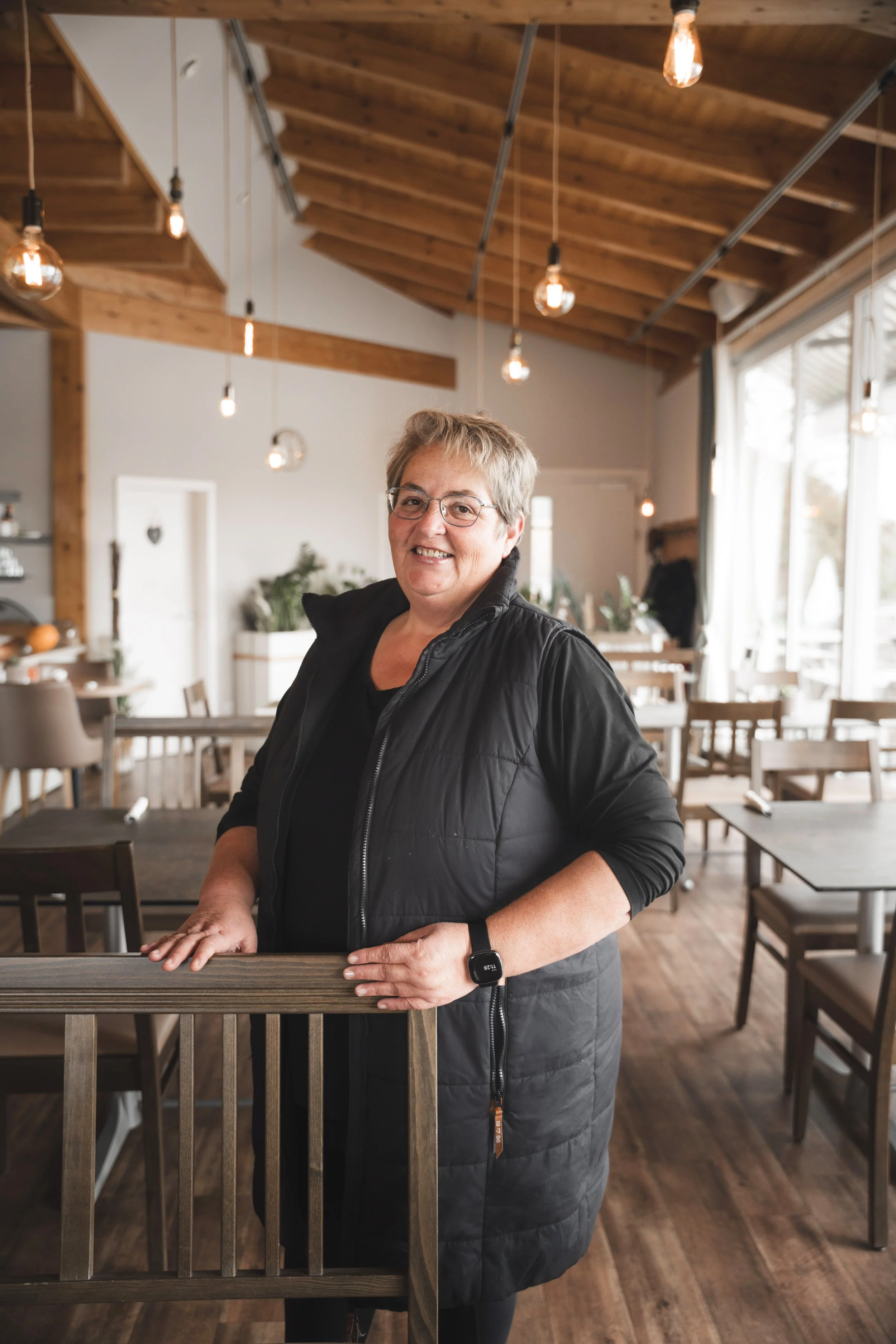 Lächelnde ältere Frau in Schwarz, die in einem gemütlichen Restaurant Posieren macht, mit Daumen und Peace-Zeichen, trägt eine schwarze Jacke, Brille und Smartwatch, im Hintergrund helle Holzdecke und Lampen.
