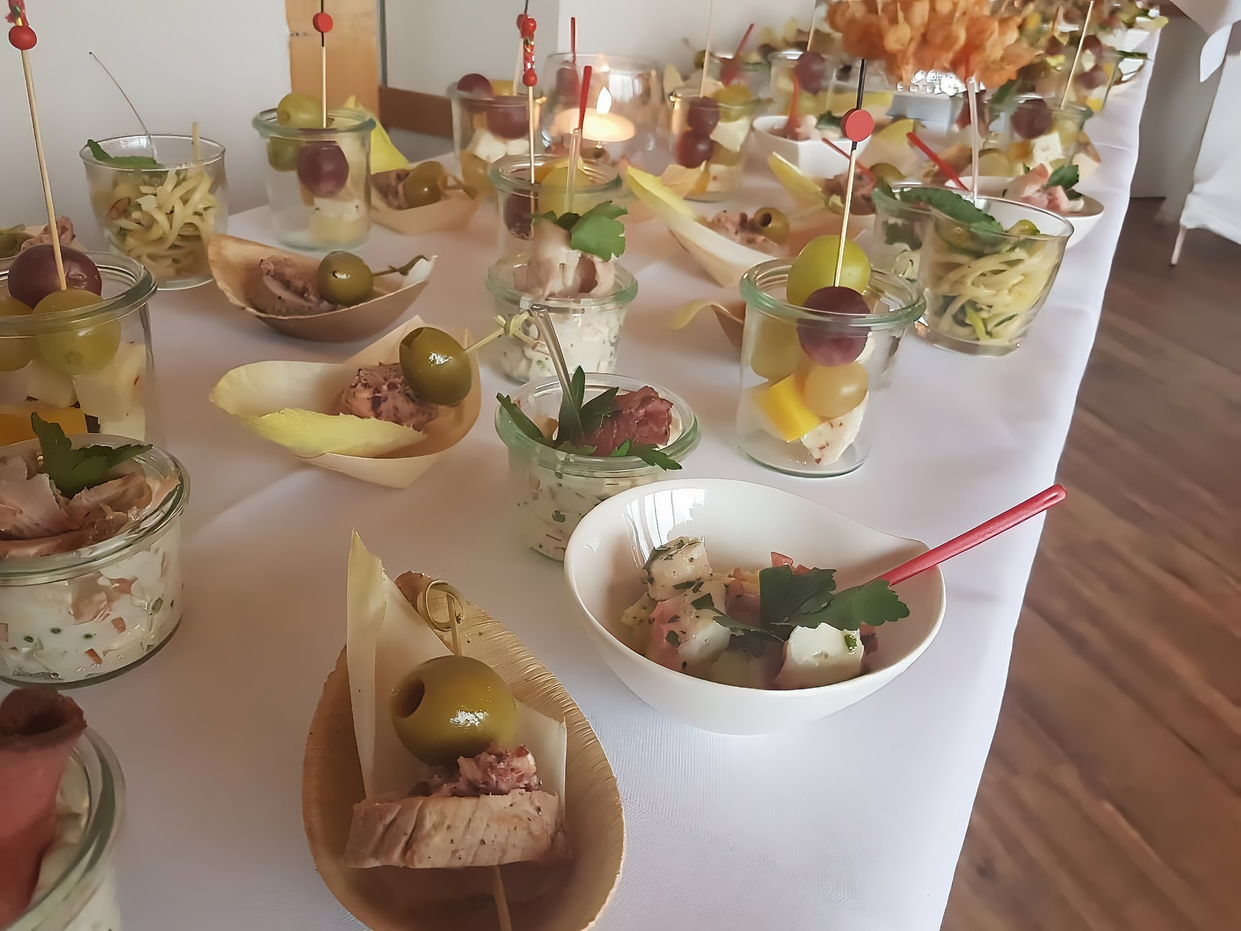 Fingerfood im Haus am See: Verschiedene kleine Gläser und Schälchen mit Vorspeisen, darunter Käse, Aufschnitt, Weintrauben und Oliven, auf einem weißen Tischtuch bei einer Feier.