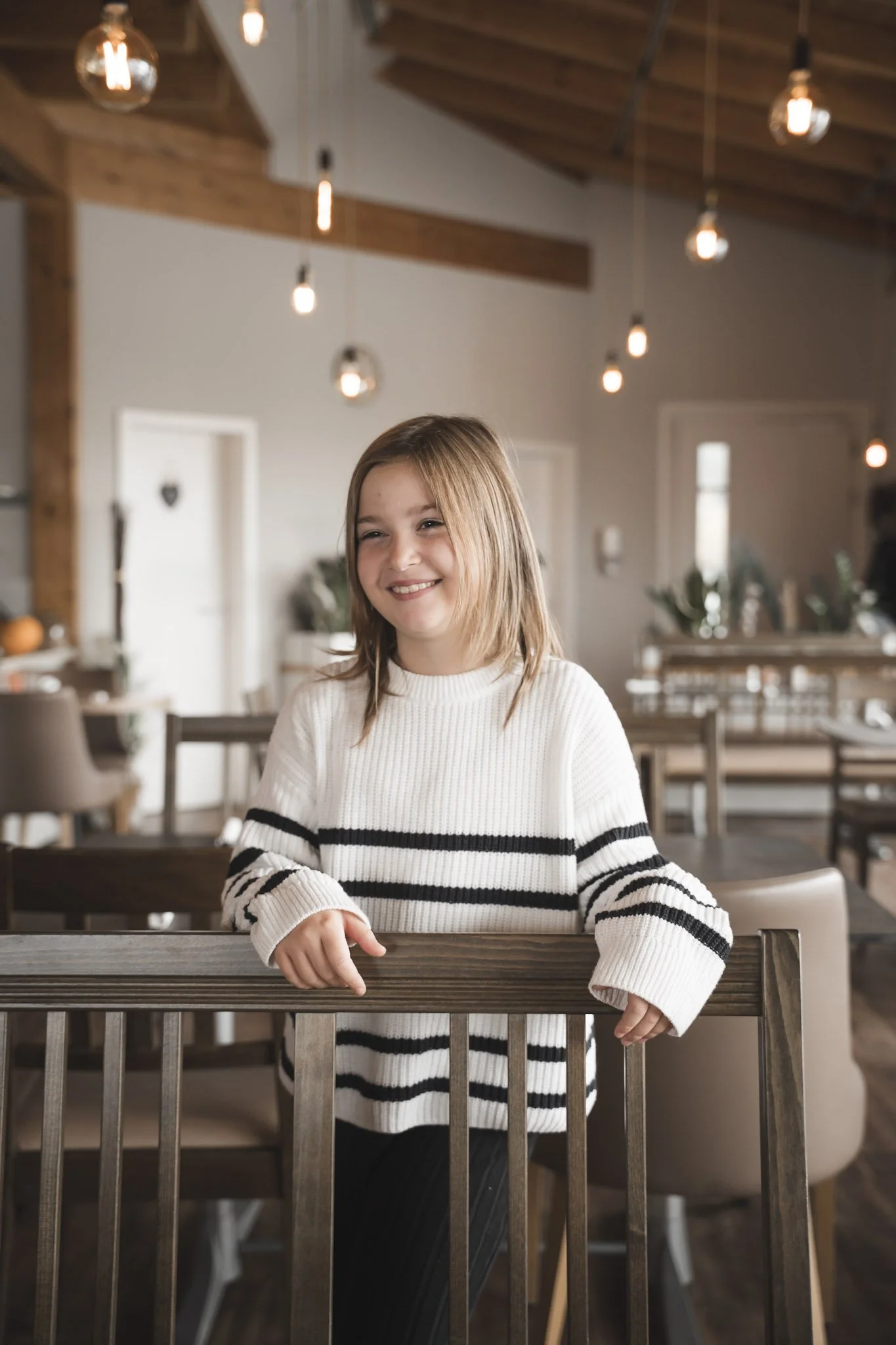 Ein lächelndes Mädchen in einem weißen Pullover mit schwarzen Streifen, das an einem Holztisch im Restaurant sitzt.