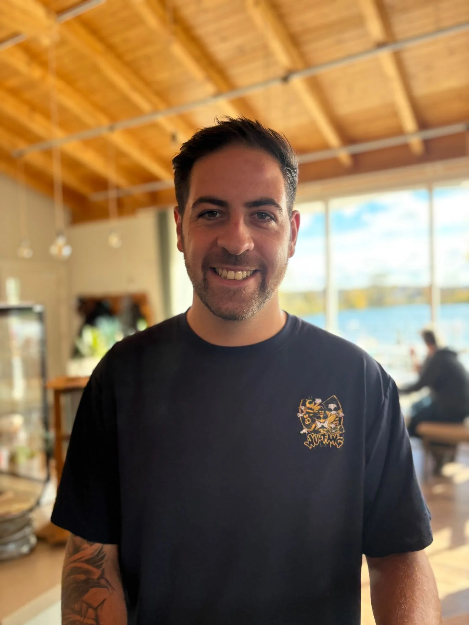 Ein lachender junger Mann mit dunklem Haar und Bart steht in einem hellen Raum mit Holzdach und großen Fenstern, im Hintergrund sind andere Personen zu sehen.