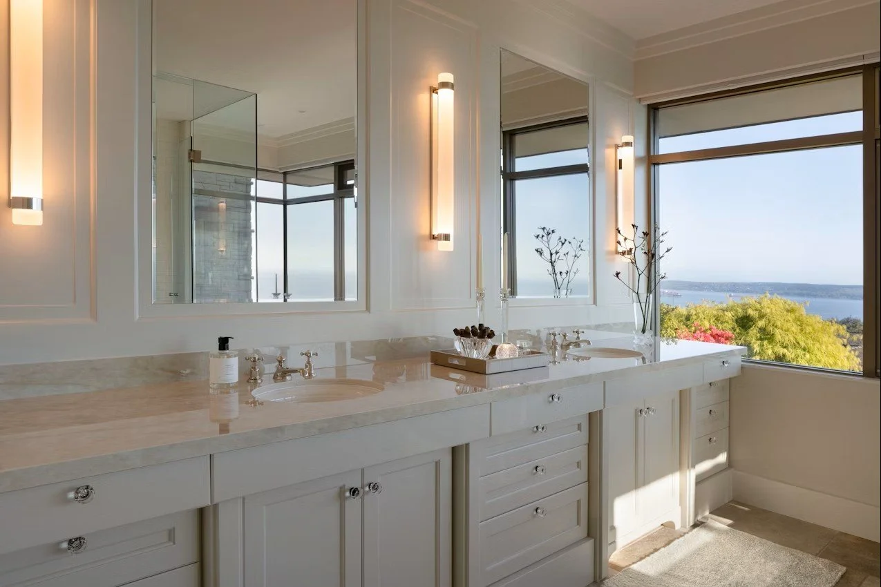 Salle de bain avec double lavabo, grands miroirs, murs blancs, vue sur la mer et la nature par de grandes fenêtres, éclairage mural
