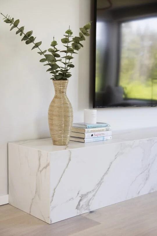 Vase en osier avec des branches de eucalyptus sur une console en marbre blanc, à côté d'une pile de livres et d'une télévision à écran plat.