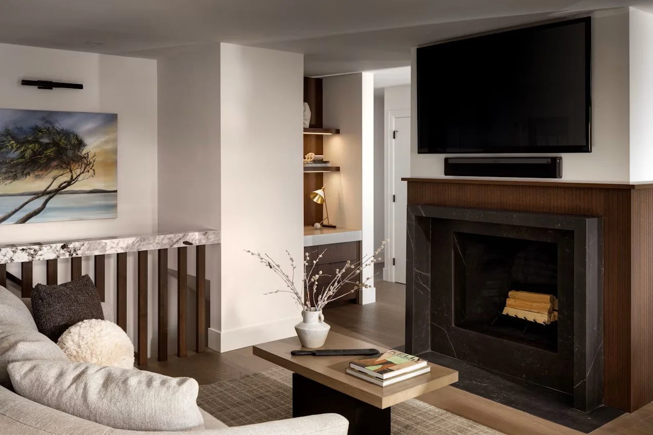 Salon moderne avec cheminée en marbre noir, télévision à écran plat, canapé beige avec coussins, tableau de paysage au mur, table basse avec livres et vase avec branches, étagère en bois avec lampe