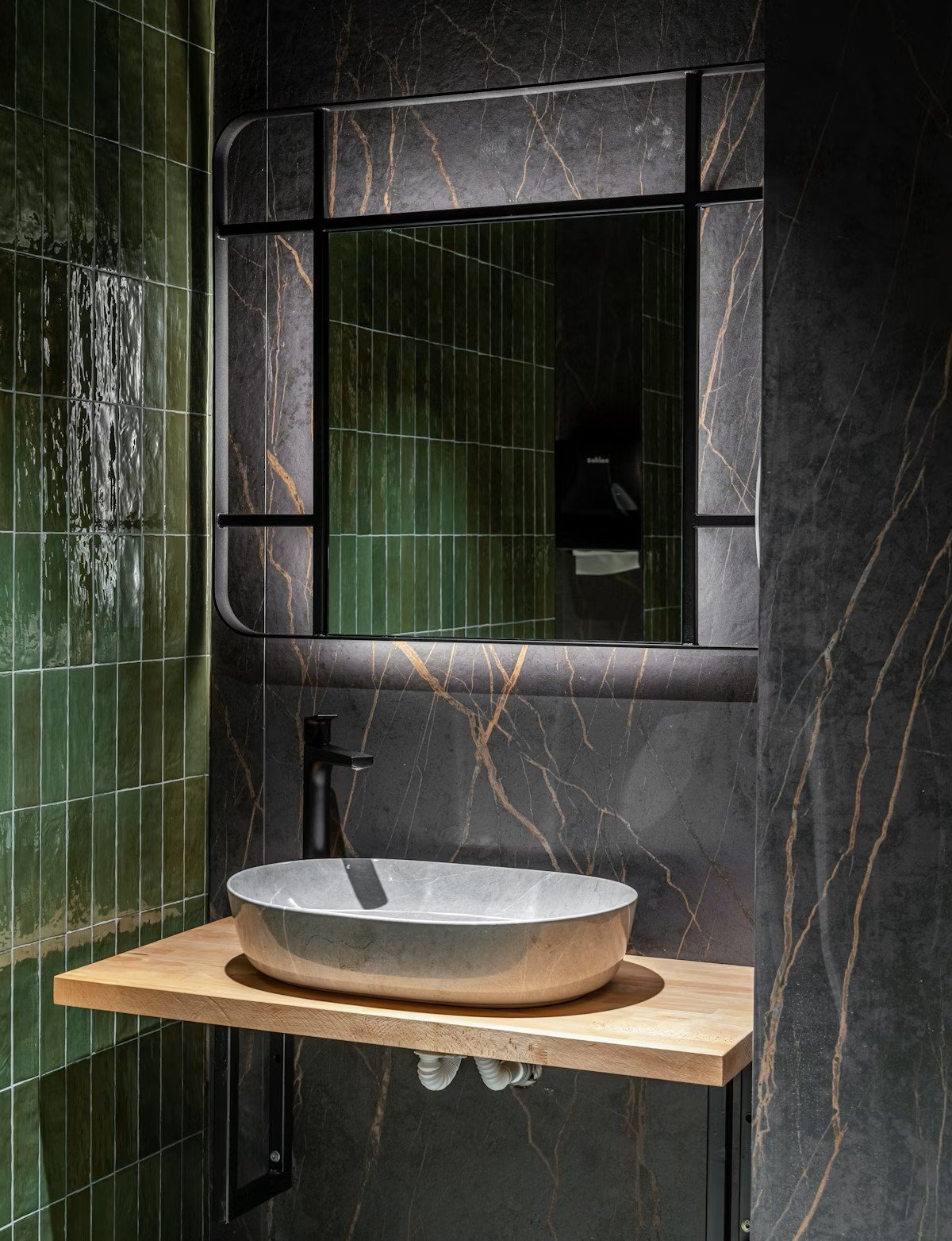 Lavabo en pierre grise oval avec un robinet noir posé sur une étagère en bois, miroir noir encadré avec éclairage intégré, murs en carreaux de marbre vert foncé et noir avec veines dorées, dans une salle de bain moderne.