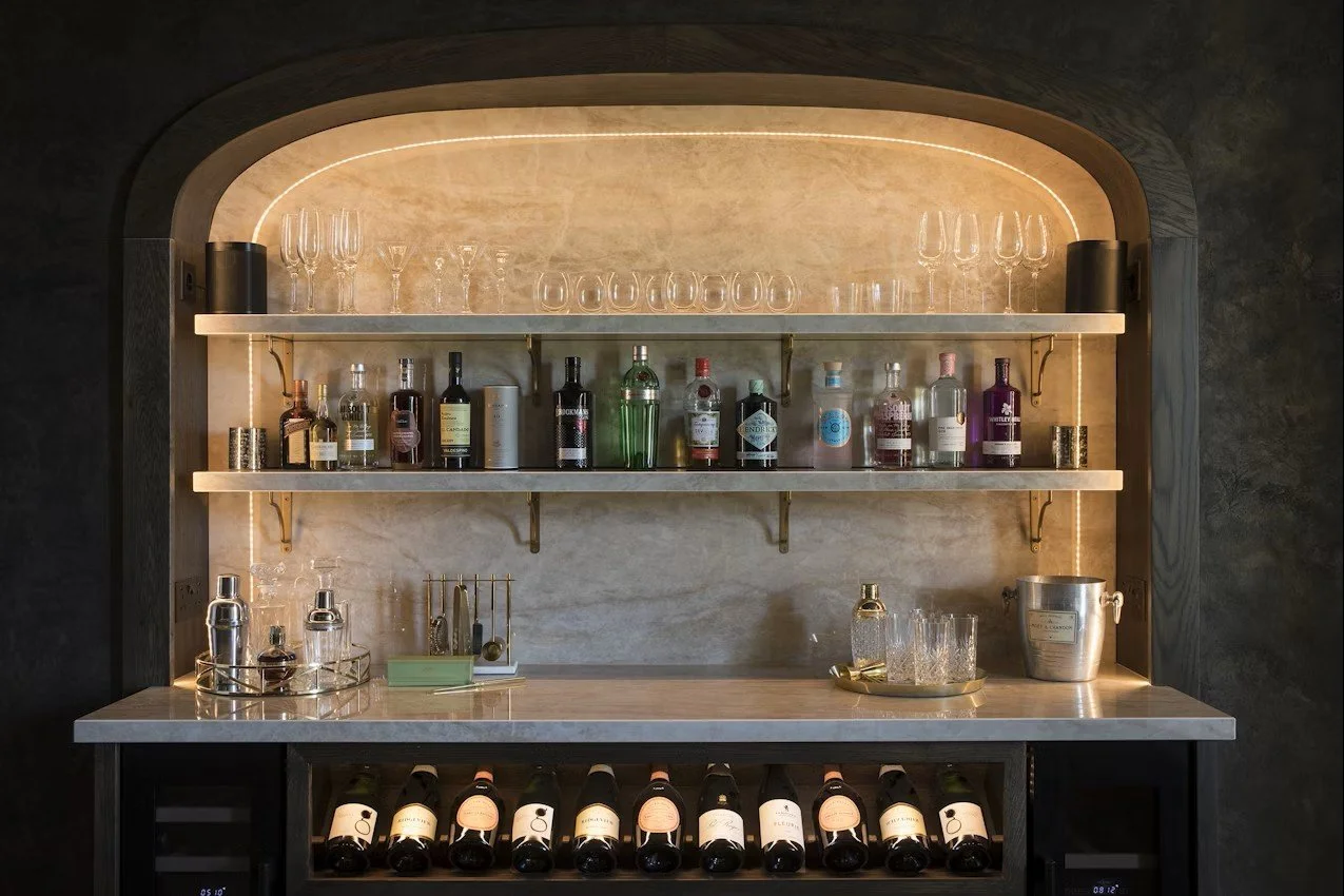 Un bar avec des étagères contenant des bouteilles d'alcool et des verres, éclairé par un éclairage intégré, avec une surface de comptoir en pierre et des bouteilles de vin rangées en dessous.