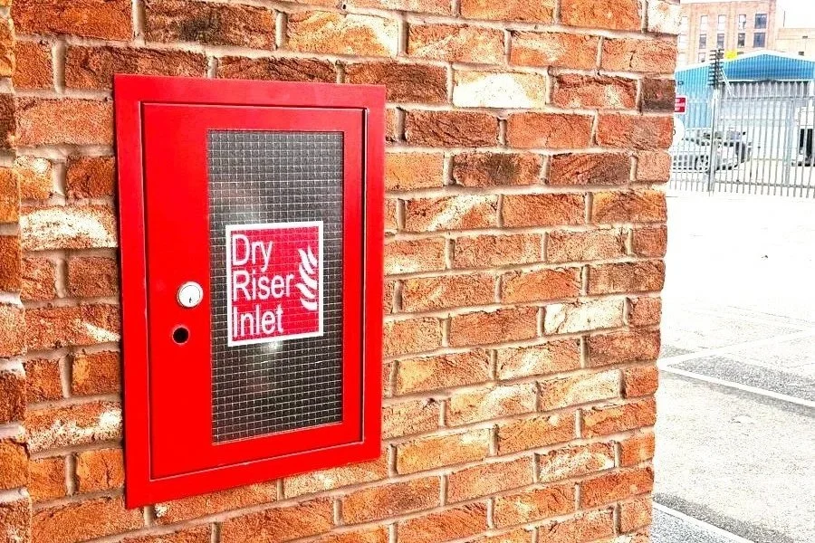 Red fire safety cabinet on brick wall labeled 'Dry Riser Inlet' with a window and a lock.