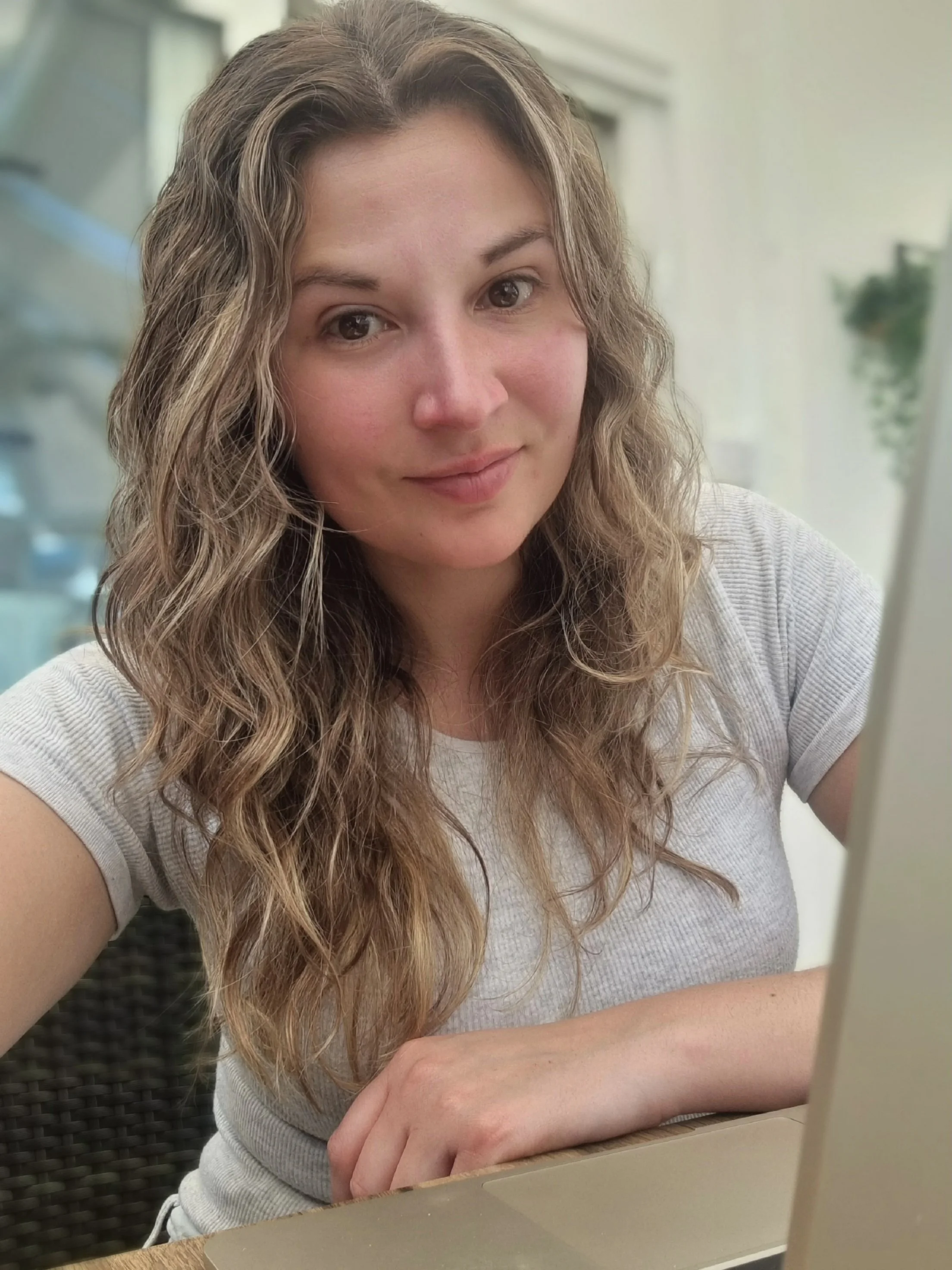A woman with long, curly hair sitting at a table indoors, looking at the camera with a slight smile.