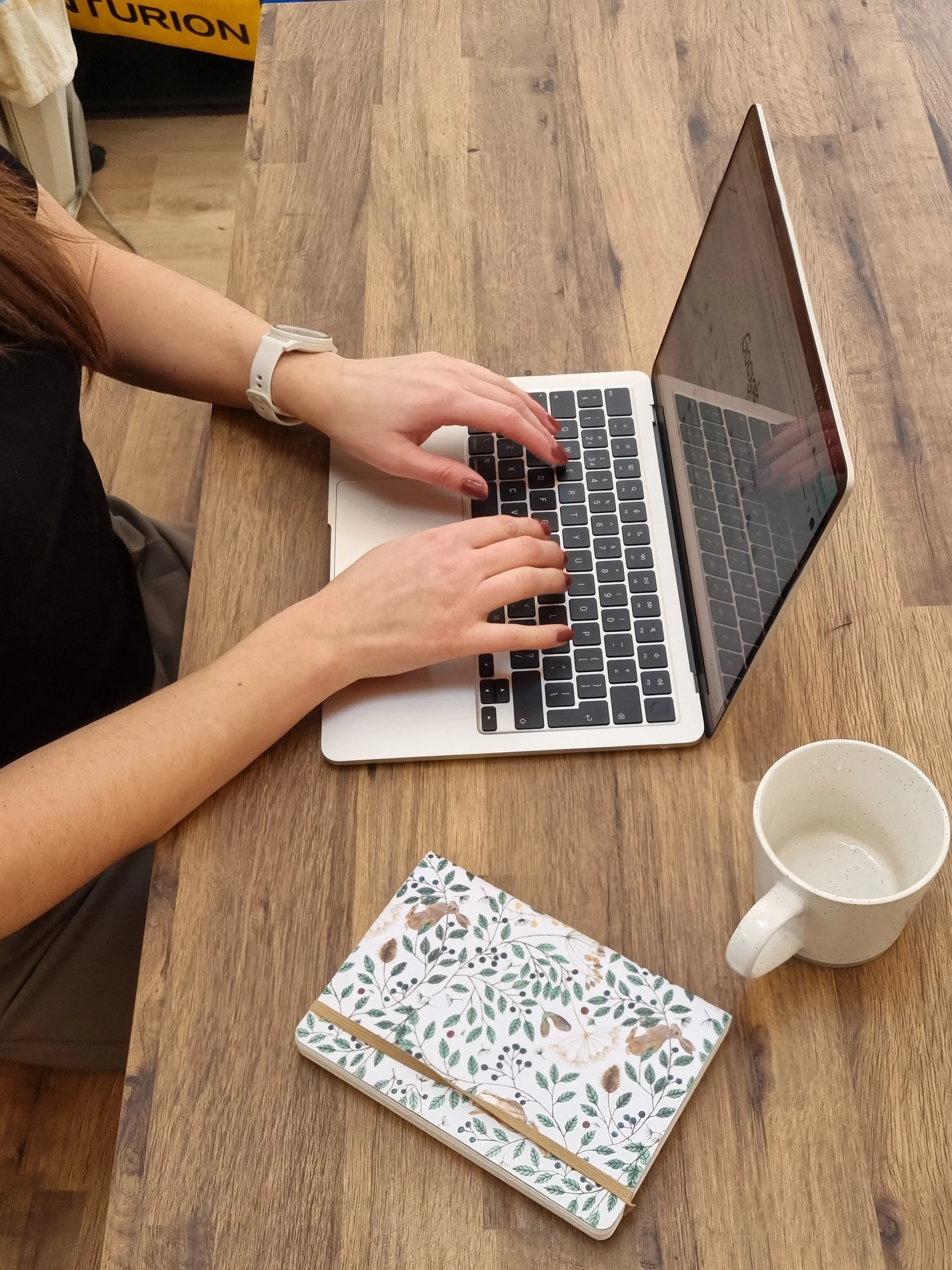 Person using a MacBook laptop at a wooden table, wearing a white watch, with a ceramic mug and botanical notebook nearby.