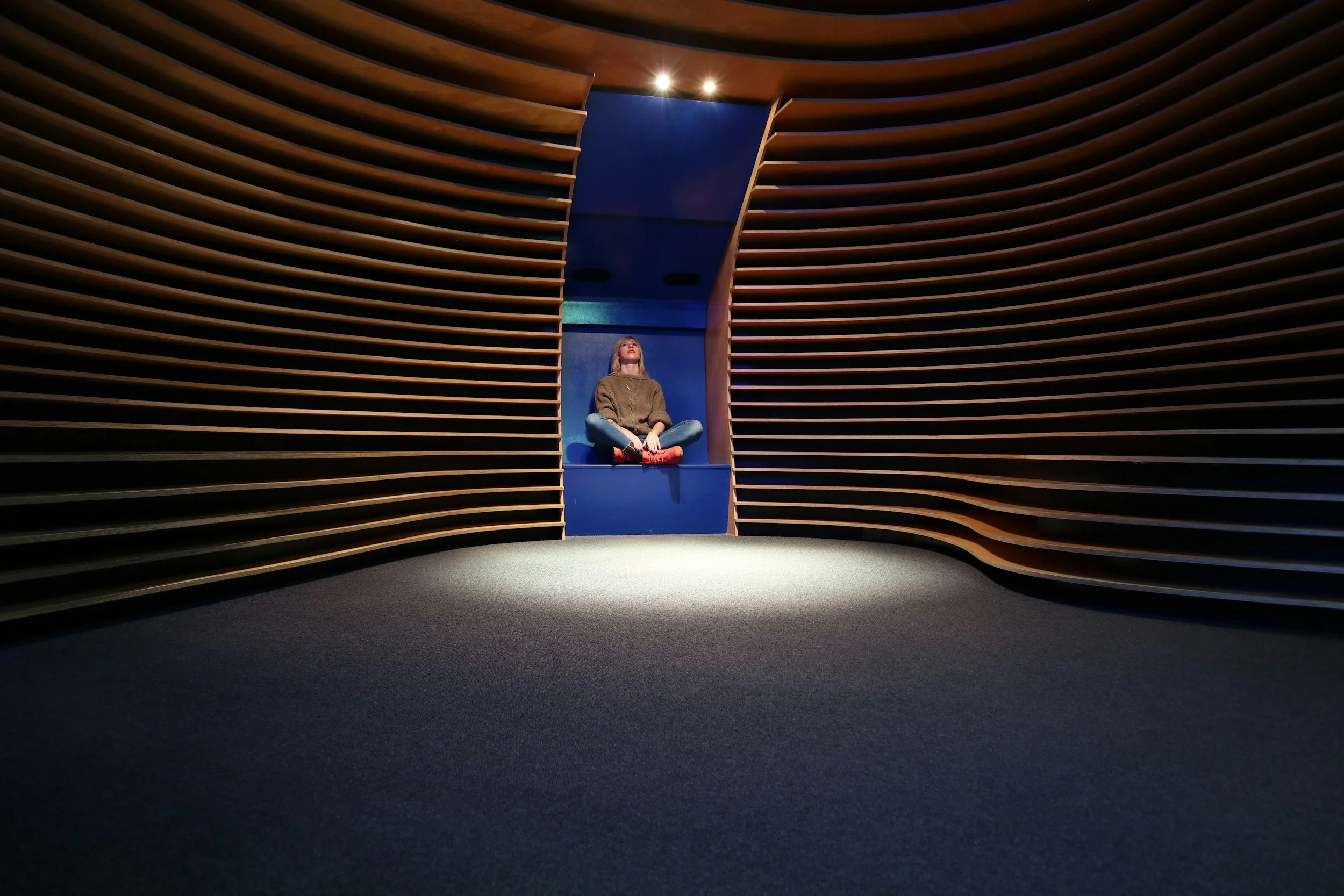 A woman sitting cross-legged on a blue ledge in a modern, curved wooden architectural space, looking upward.