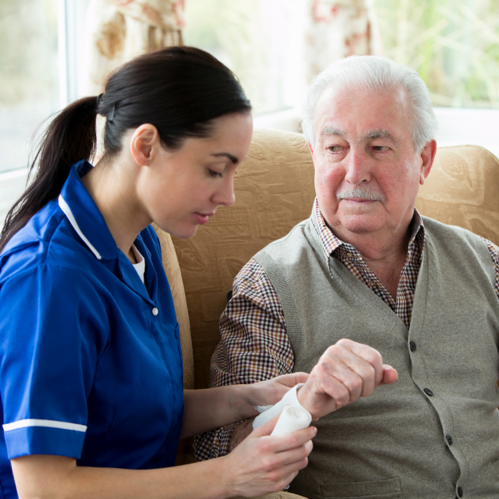 A nurse wraps a bandage around an elderly man's arm in a living room with sunlight coming through the window.