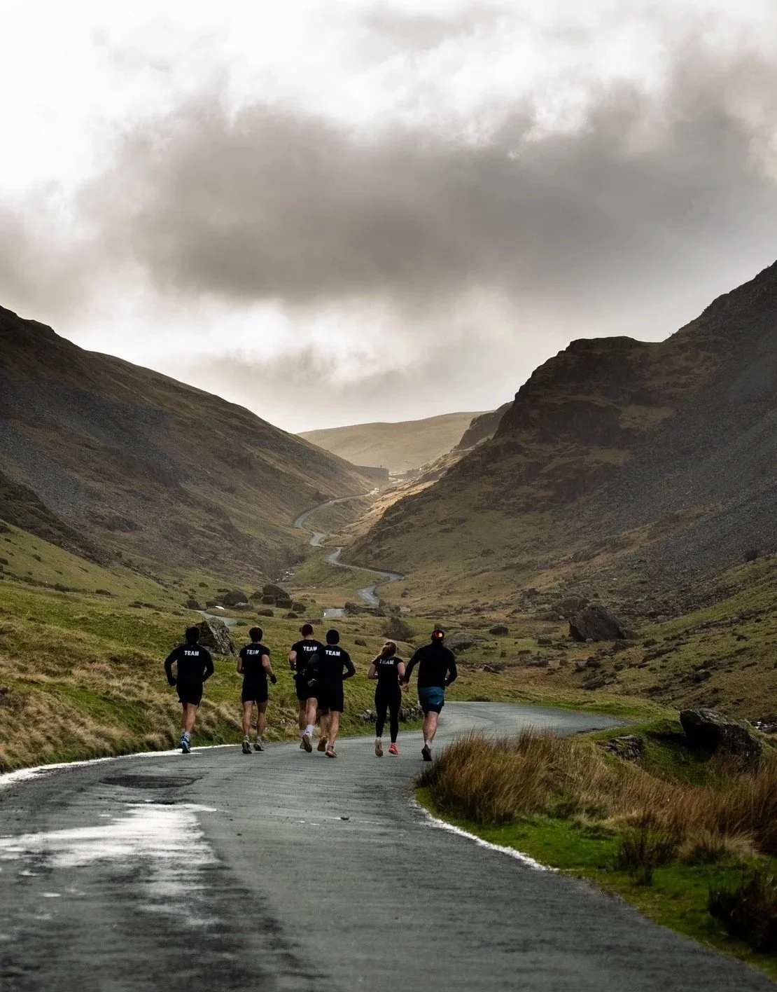 Group of six runners running on a winding mountain road in a scenic valley with green hills and cloudy sky.