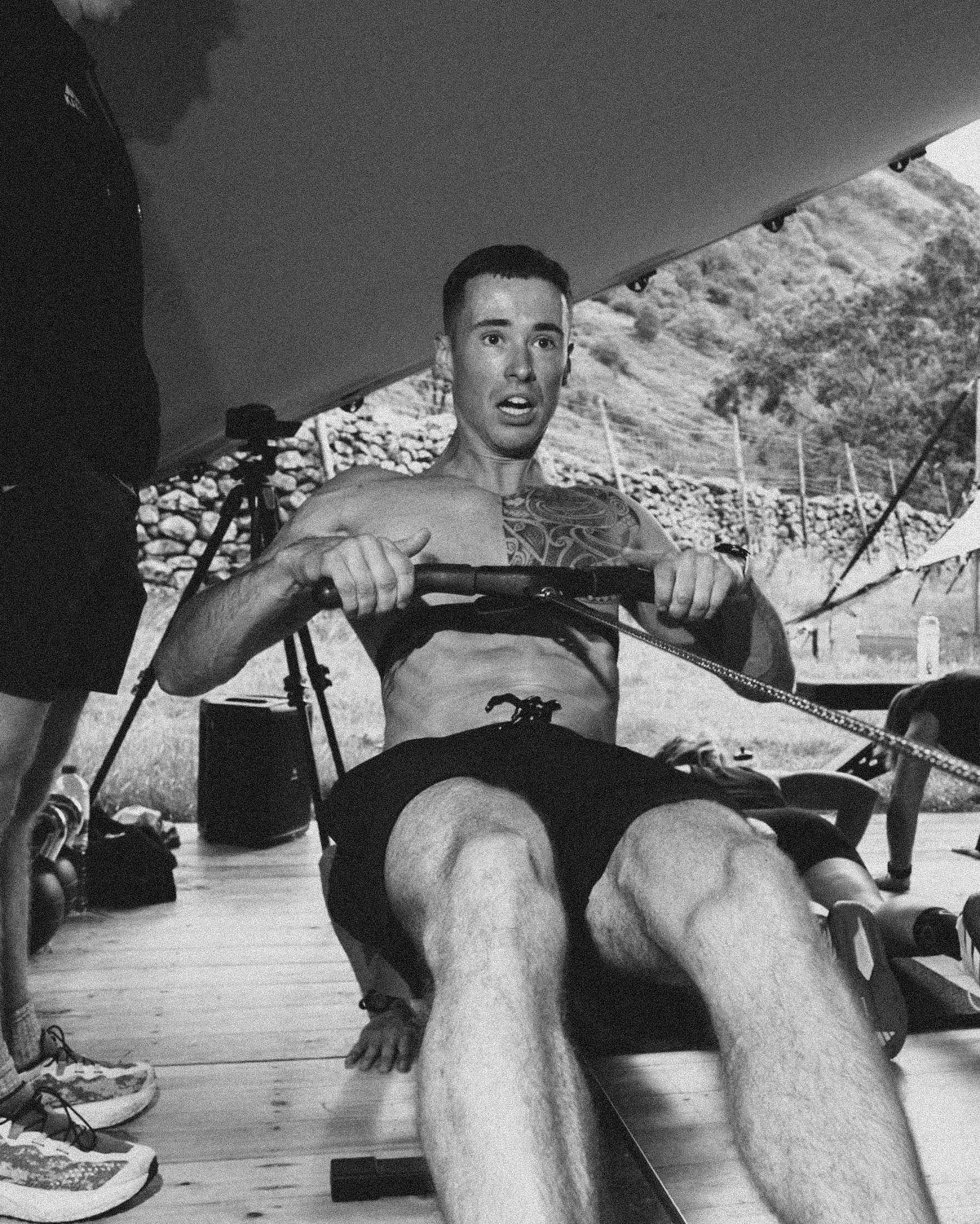 A man with tattoos on his chest and arm, wearing shorts, is sitting on a rowing machine, looking surprised or focused, indoors at a gym or training facility.