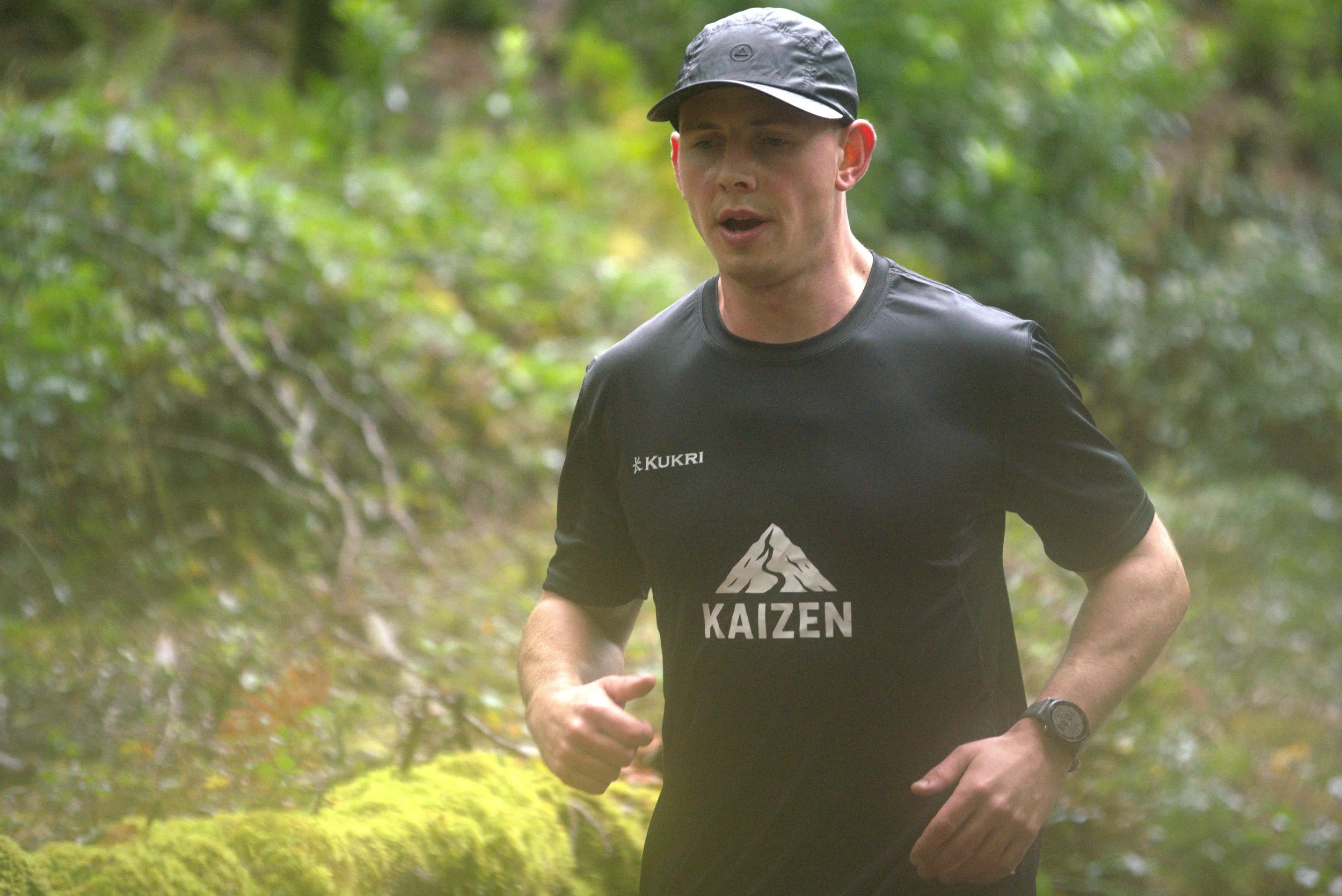 A shirtless man with a tattoo on his back running on a track indoors, with other runners in the background.