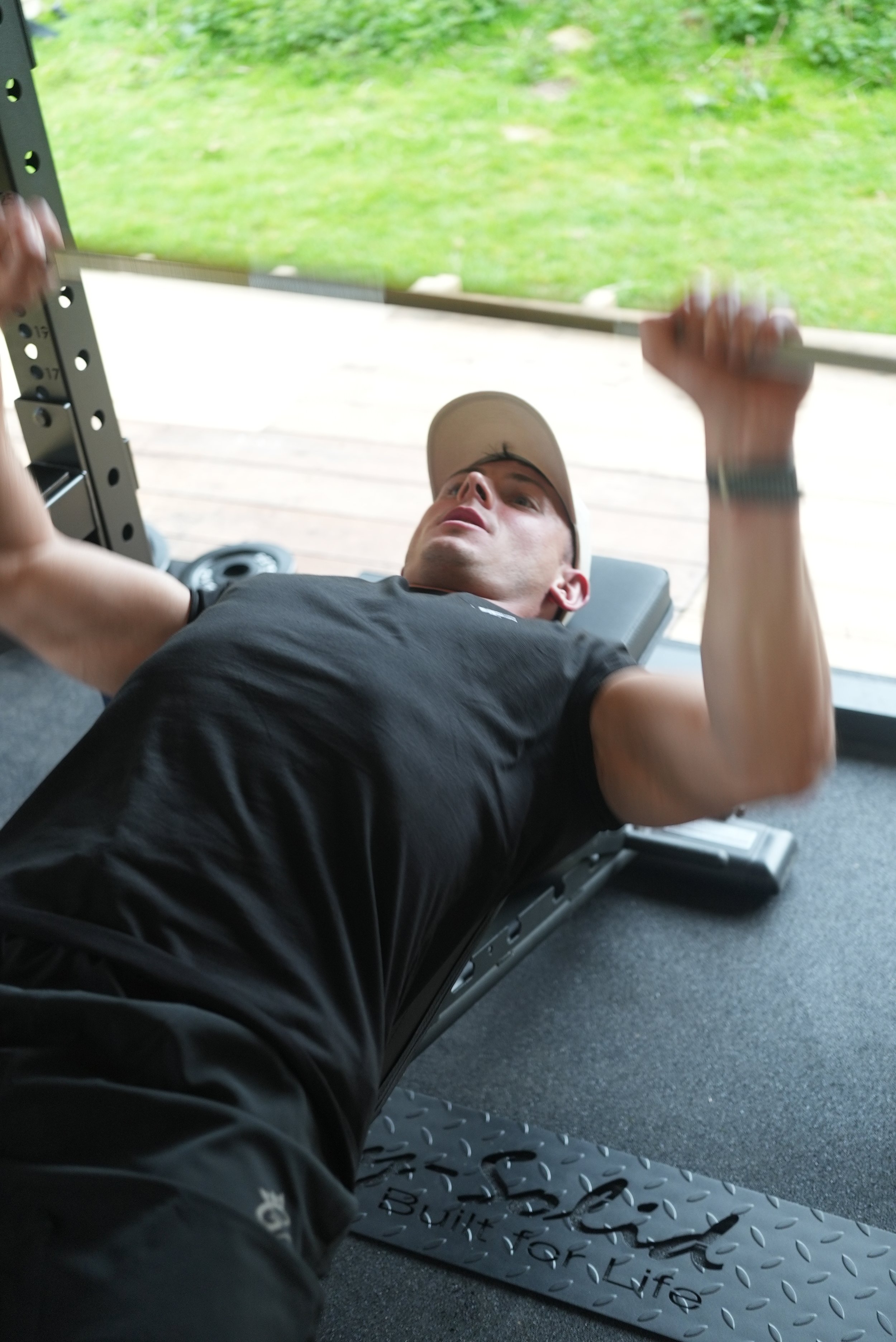 Close-up of a muscular person wearing a black shirt with the words "KAIZEN" and a mountain logo, in a gym or workout environment.