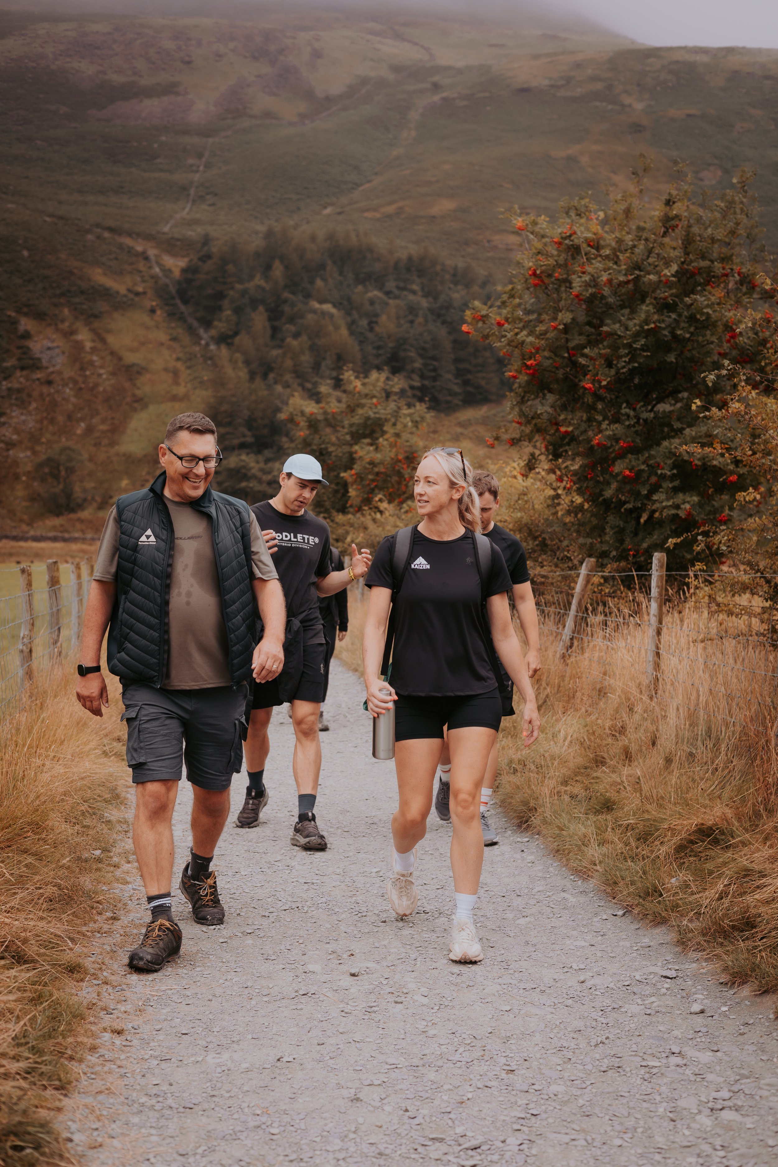 Group of people walking on a trail in a hilly, rural area during daytime. They are dressed in casual outdoor attire, with some smiling and engaged in conversation.