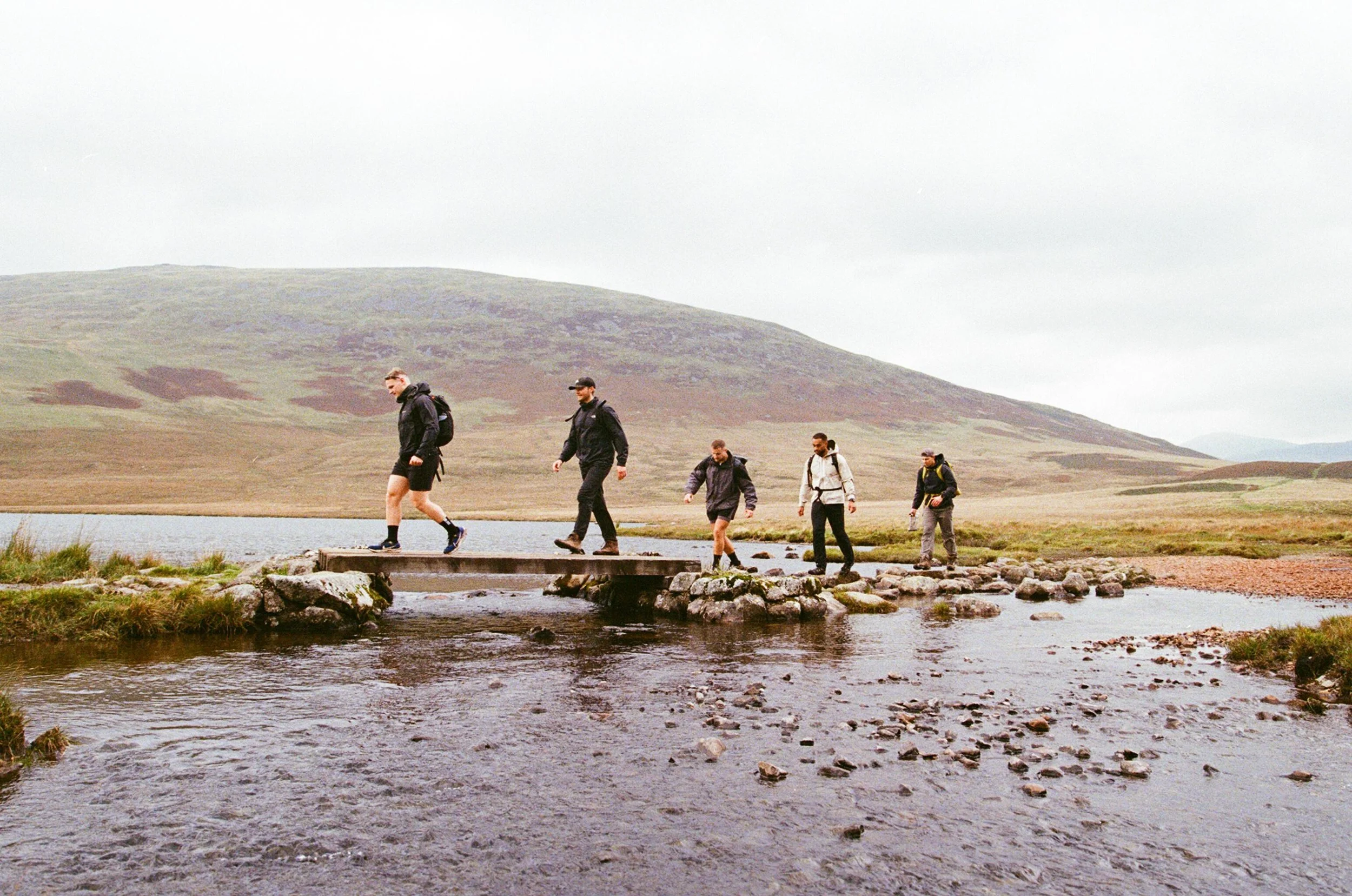 Young man wearing a dark cap and blue shirt with a black backpack, standing outdoors in a hilly area with a cloudy sky.