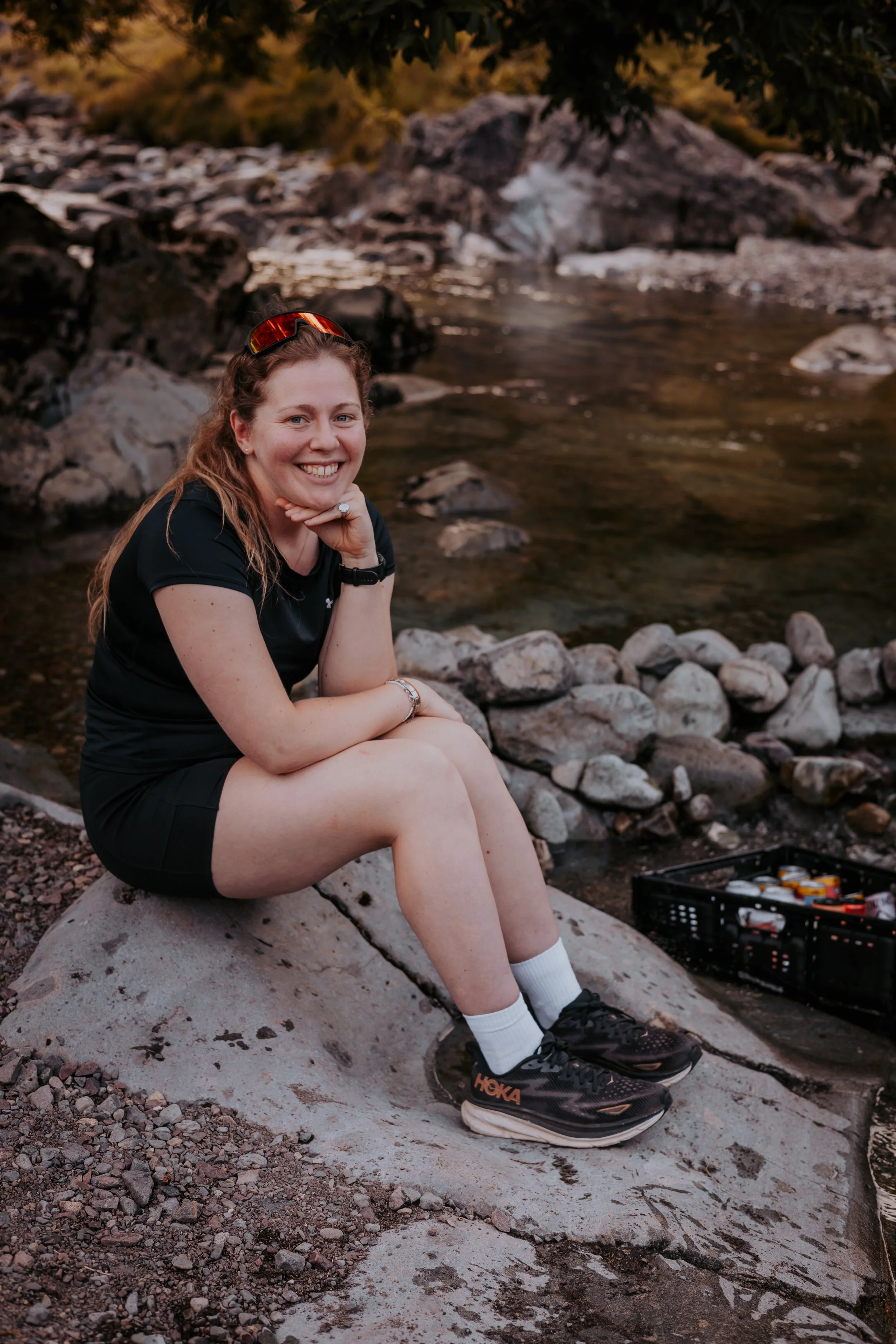 A woman sitting on a large rock near a riverbank, smiling at the camera, with a tool kit and camping gear nearby, surrounded by rocks and trees.