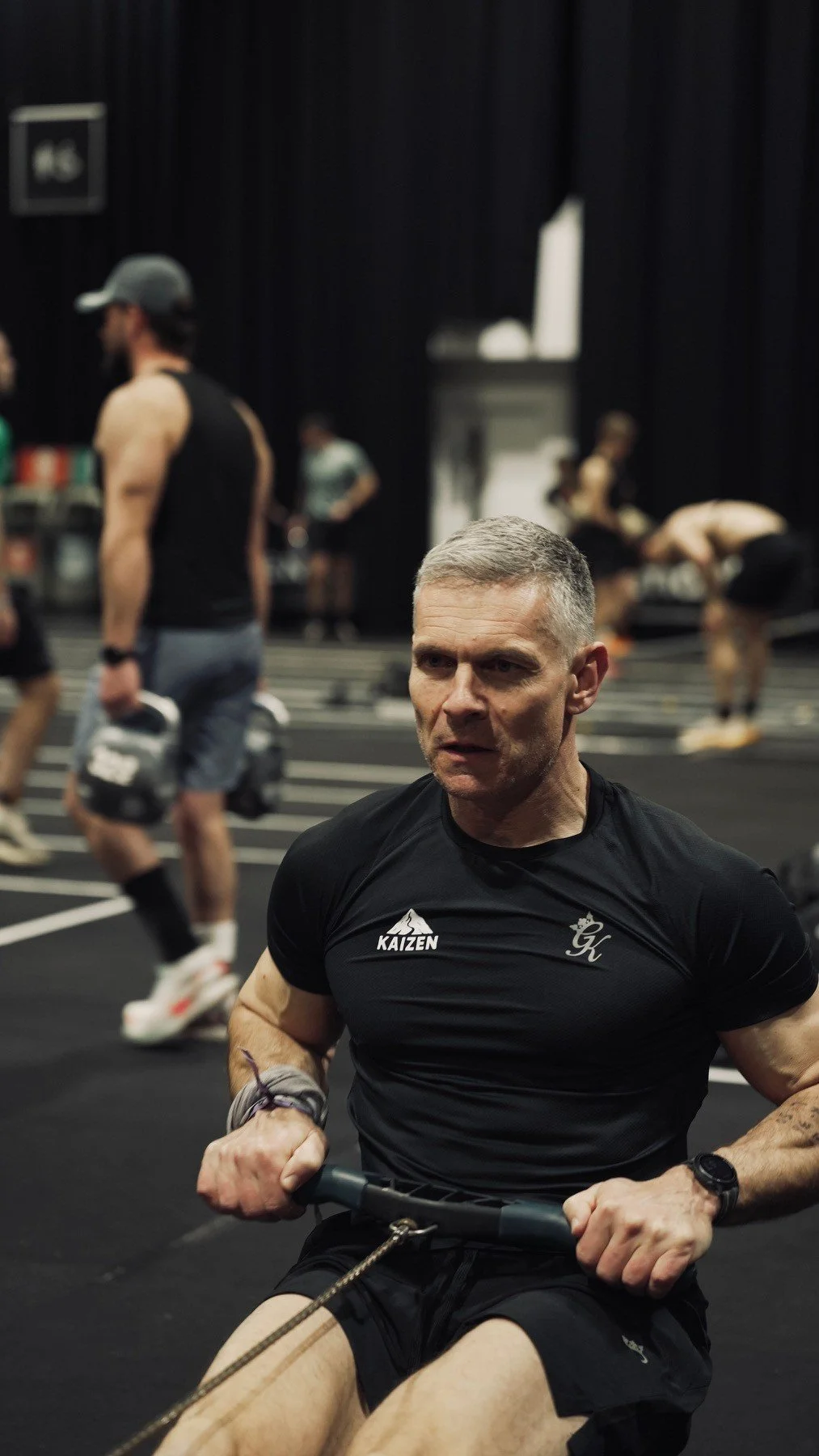 A man in a black shirt and shorts is working out with a rowing machine in a gym. There are other people exercising in the background.