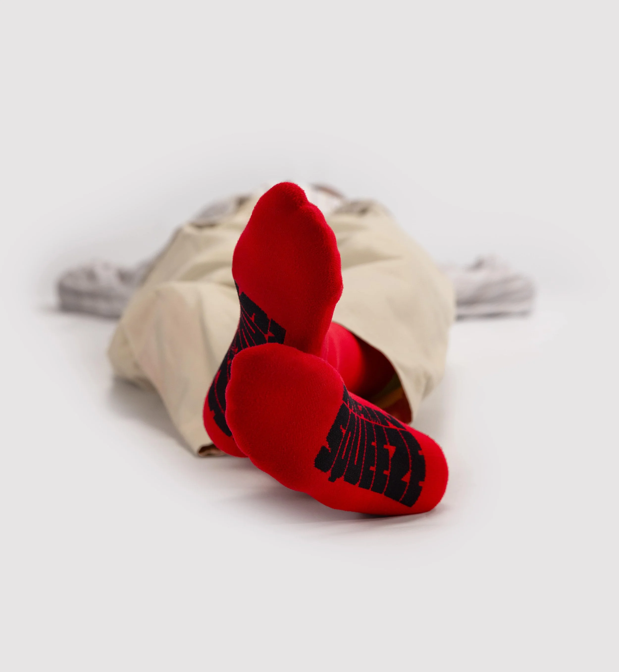 Person lying down with legs and feet raised, wearing bright red socks with black designs, beige shorts, and gray sneakers, on a white background.
