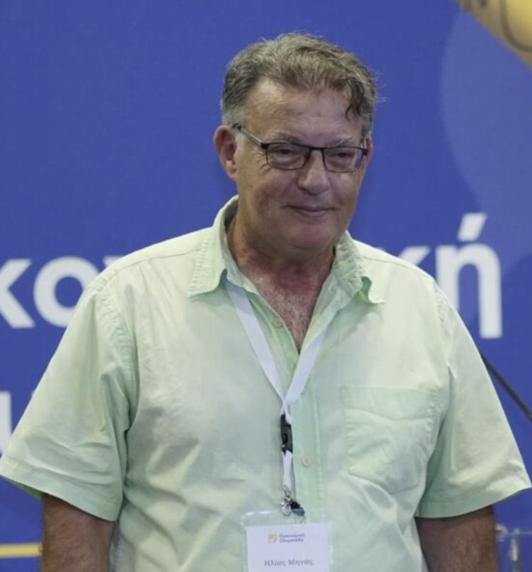Portrait of a man wearing glasses, a white shirt, and a dark blazer, standing indoors near windows with natural light.