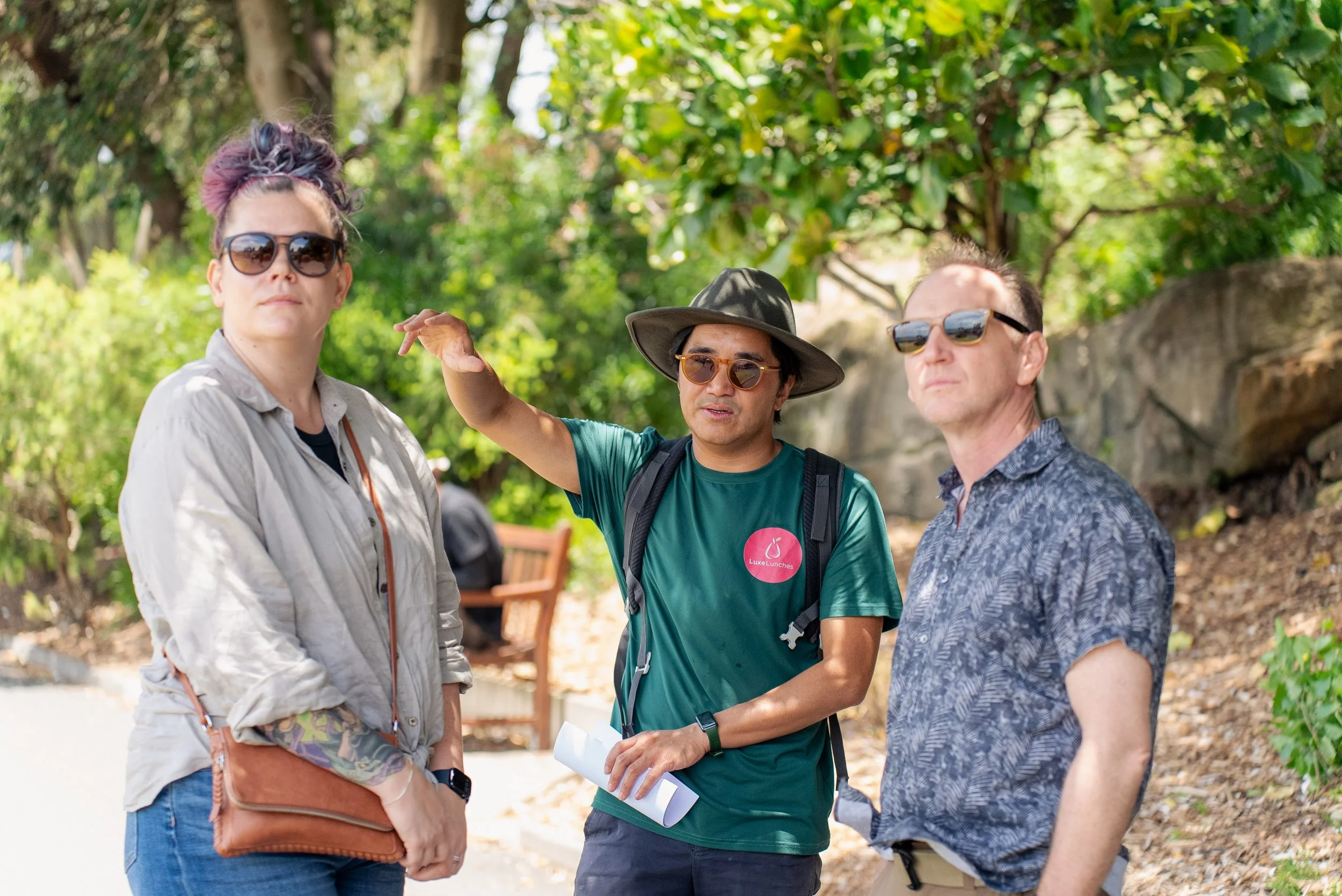 Three people outdoors on a sunny day, two men and one woman, engaged in conversation with trees and a stone wall in the background.