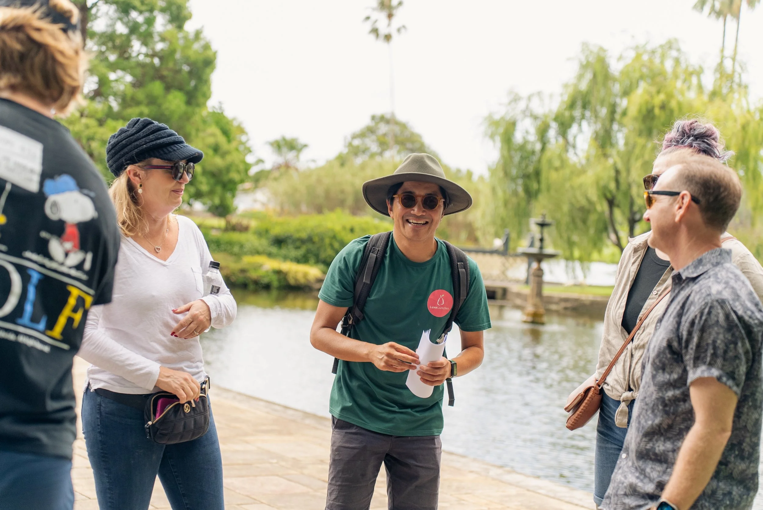 Group of five people standing outdoors near a pond, having a conversation on a cloudy day.