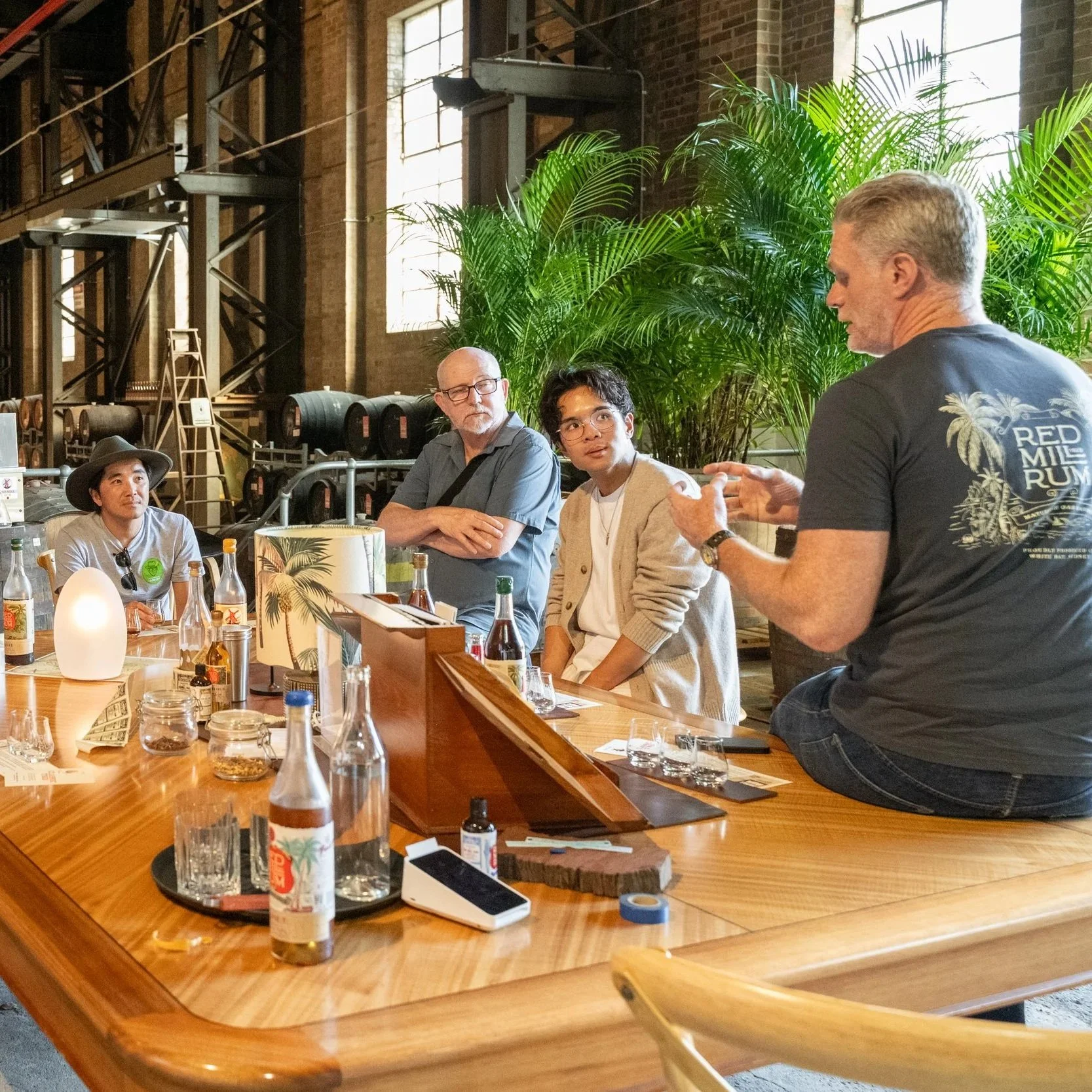 A group of four people sitting at a wooden table, listening to a man who is speaking and gesturing with his hands, inside an industrial-style room with large windows and green plants.