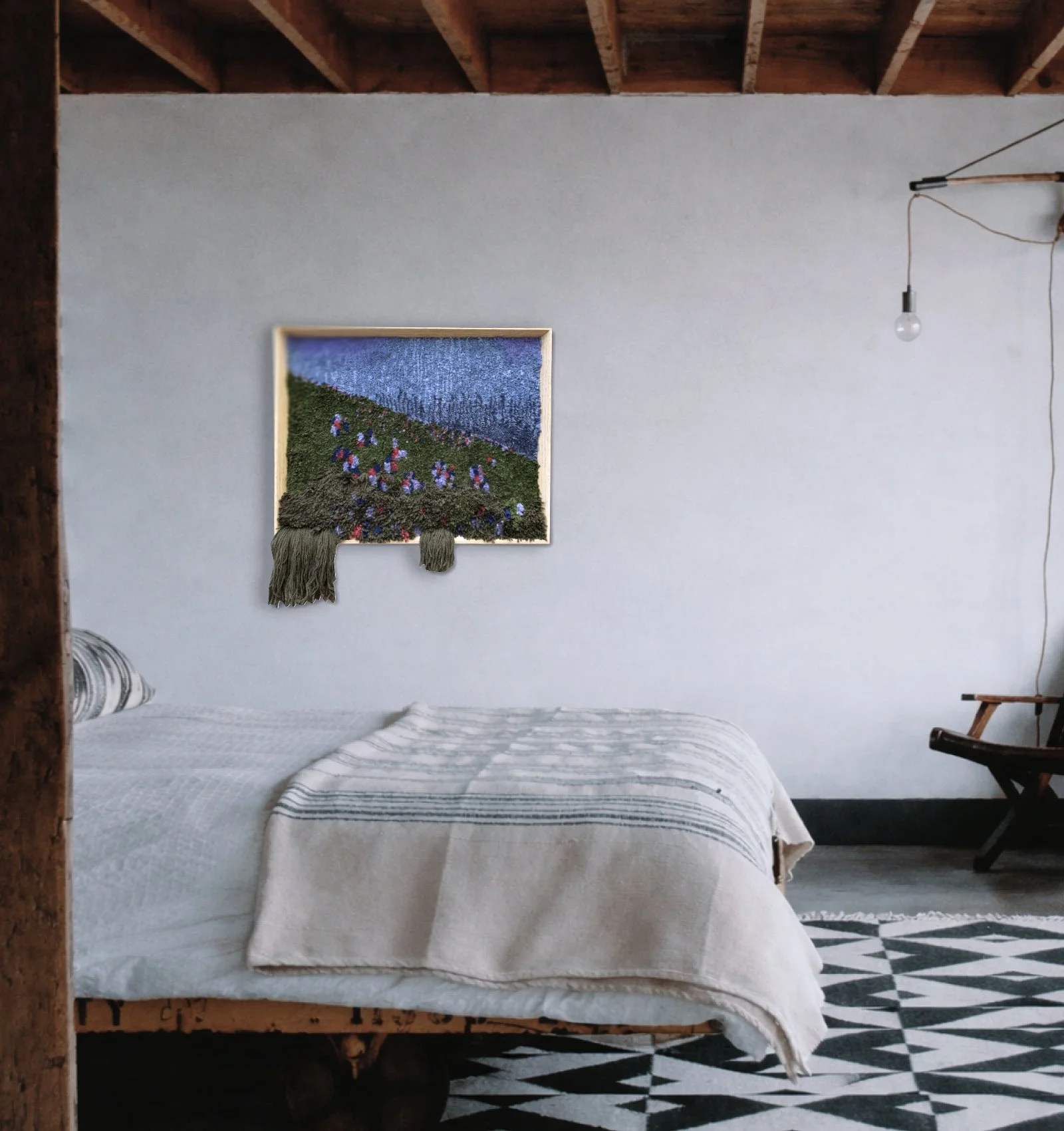 A bedroom with a bed covered in a cream-colored quilt, a striped pillow, and a wall hanging artwork of a flower field above the bed. The room has exposed wooden beams on the ceiling, a hanging light bulb, and a patterned black-and-white tile floor.