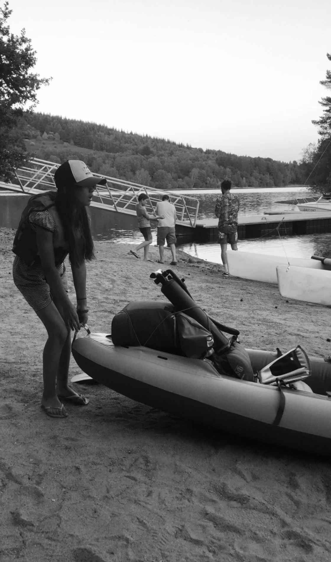People preparing for kayaking by the water, with a woman adjusting a kayak in the foreground and others near a dock in the background.