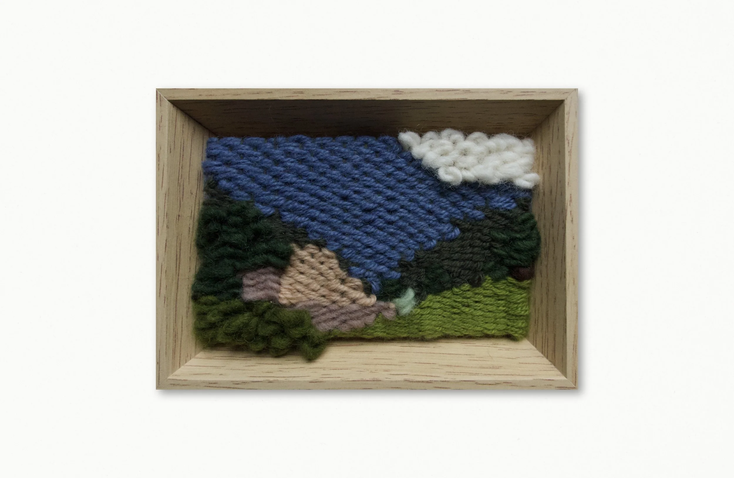 A framed textile art piece depicting a landscape scene, with a blue sky, white cloud, green hills, and a pink mountain, made from woven yarns.