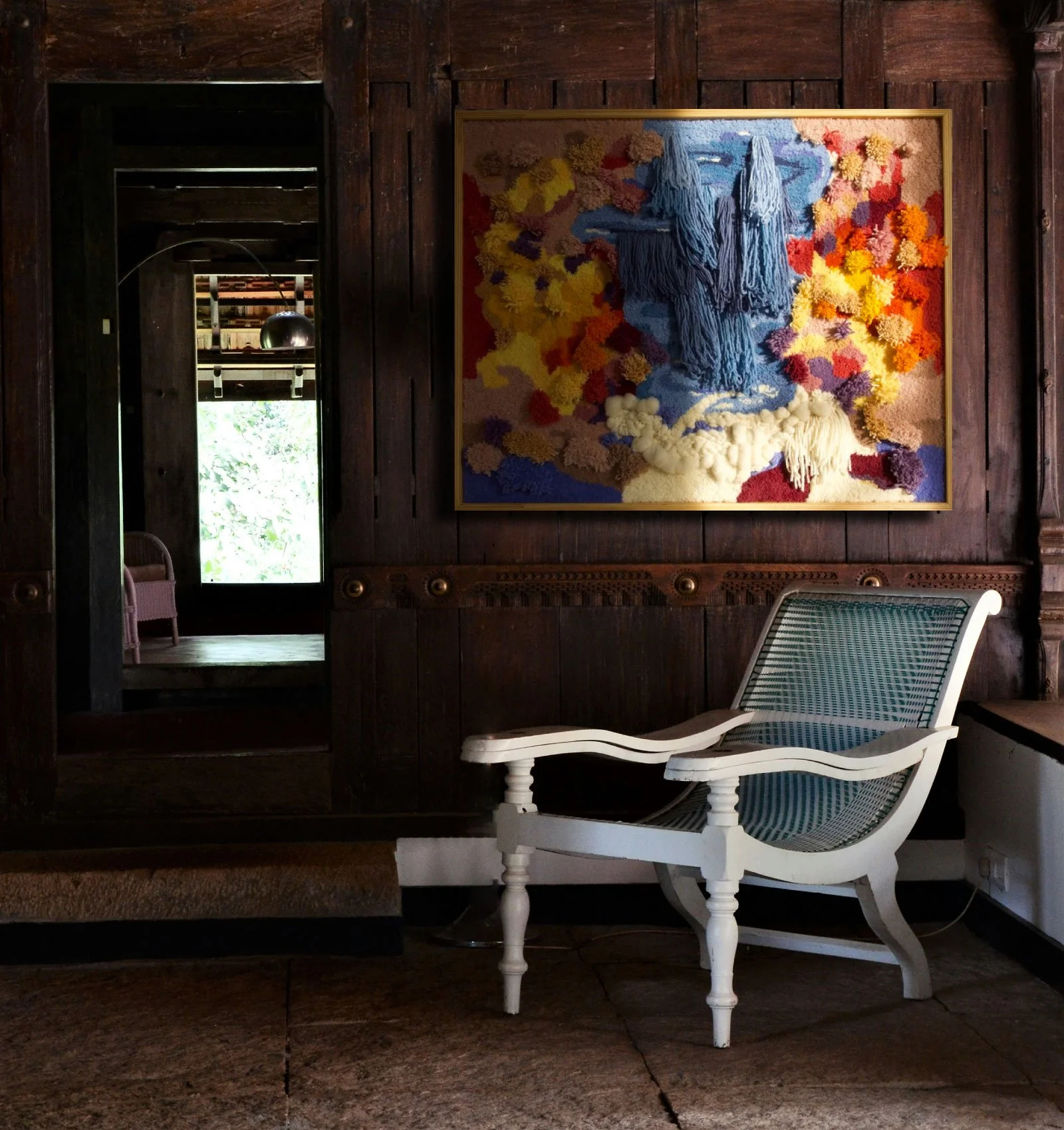 A white vintage-style chair with a blue and white checkered cushion and armrests, positioned in front of a dark wooden wall with a framed colorful textile art piece depicting a face with closed eyes and long hair made of various textured yarns and flowers.