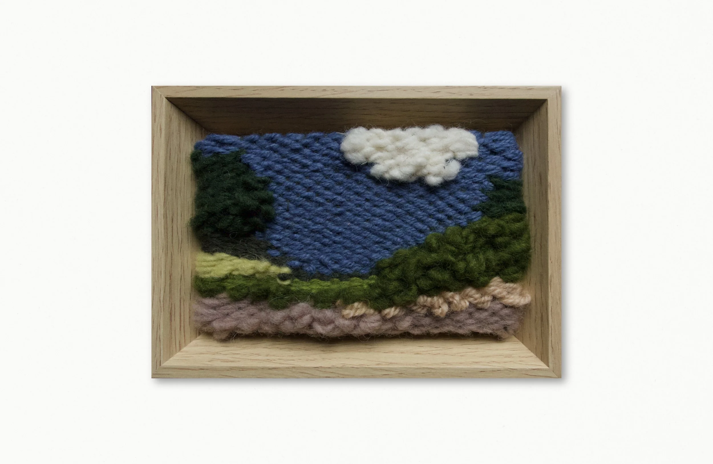 A framed artwork of a felted landscape scene with mountains, trees, blue sky, a white cloud, and ground, set in a wooden frame.