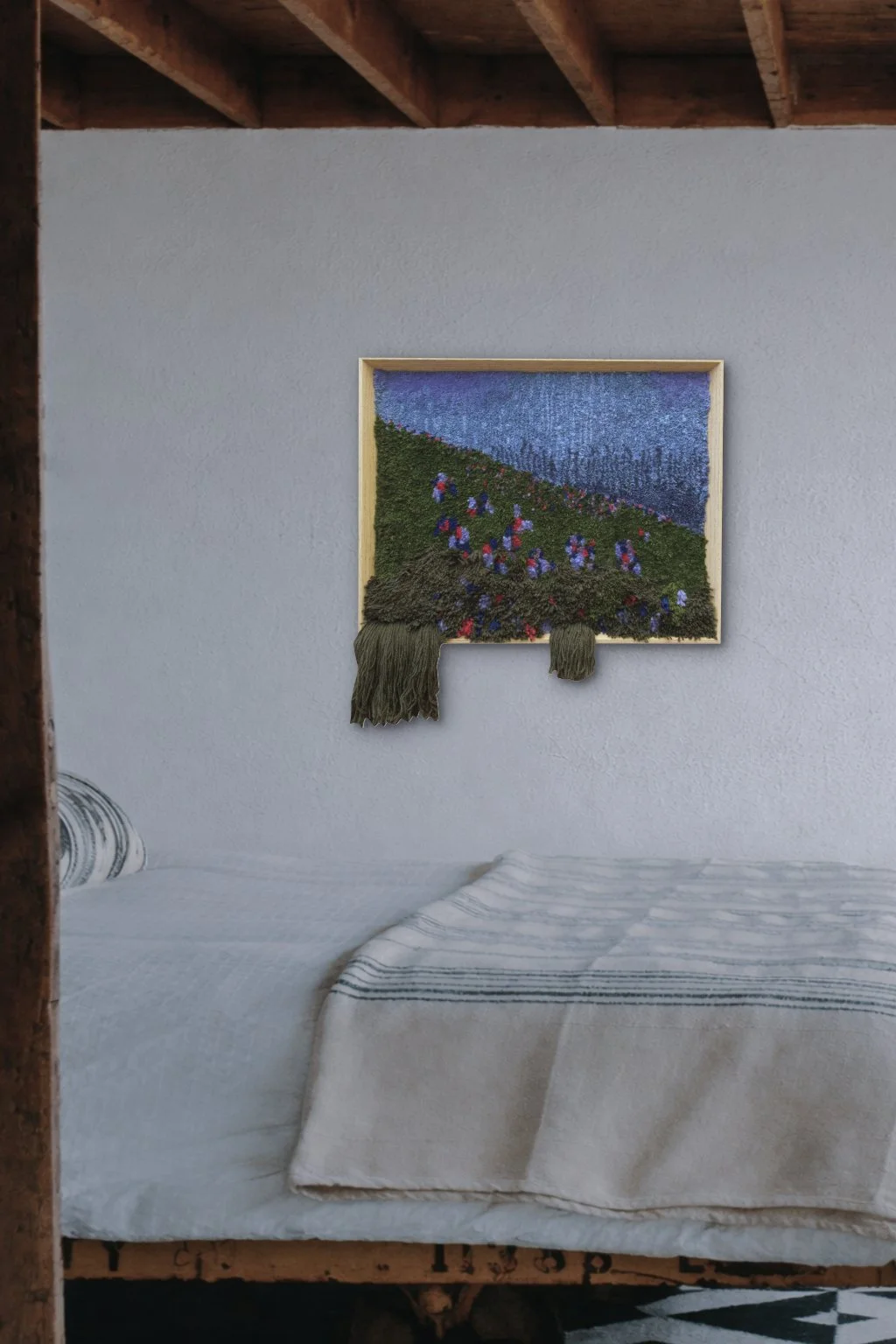 A textured woven wall hanging depicting a lavender field under a blue sky, with green grass in the foreground, hanging on a plain white wall above a bed with white and gray striped bedding.