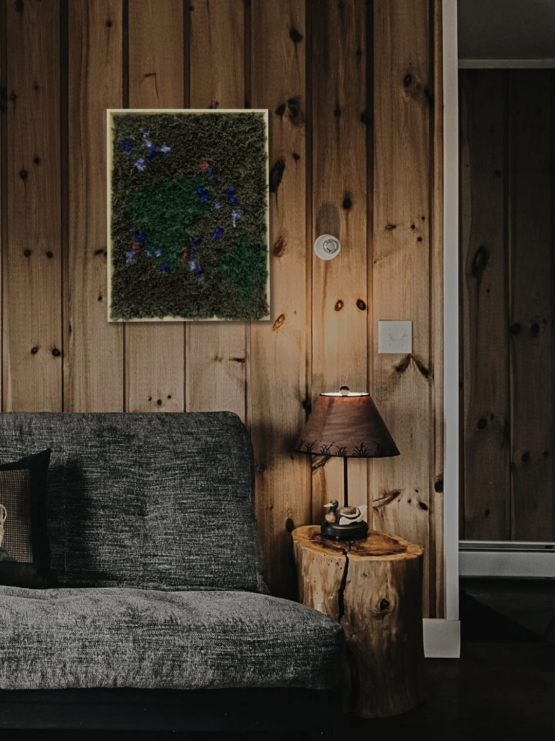 A living room corner with wood-paneled wall, framed moss art, gray upholstered sofa, and a small wooden side table with a lamp and small decorative items.