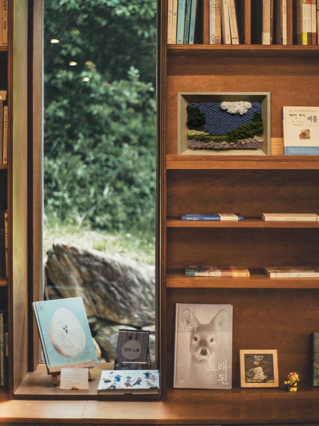 Bookshelf with books, a framed textile art piece resembling a landscape, and books on the shelves, with a large window showing greenery outside.