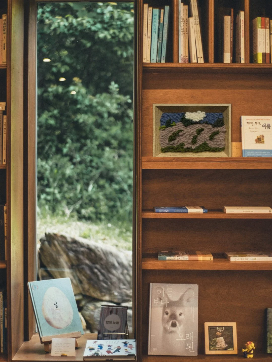 Bookshelves with books and artwork, including a framed textile art piece with mountains and clouds scene, next to a window showing greenery outside, with some books and items displayed on a window sill.