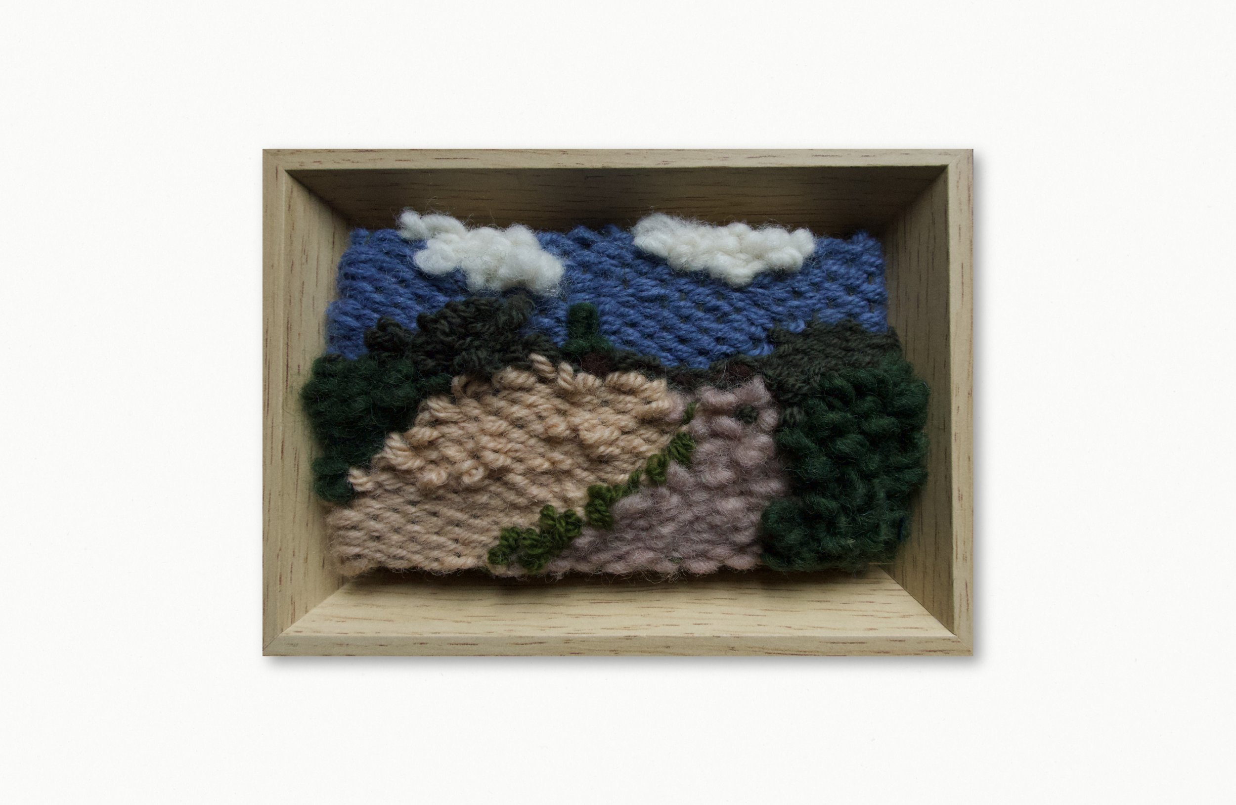 A framed textile art piece featuring a landscape scene with clouds, mountains, and greenery, crafted with thick, colorful yarn.
