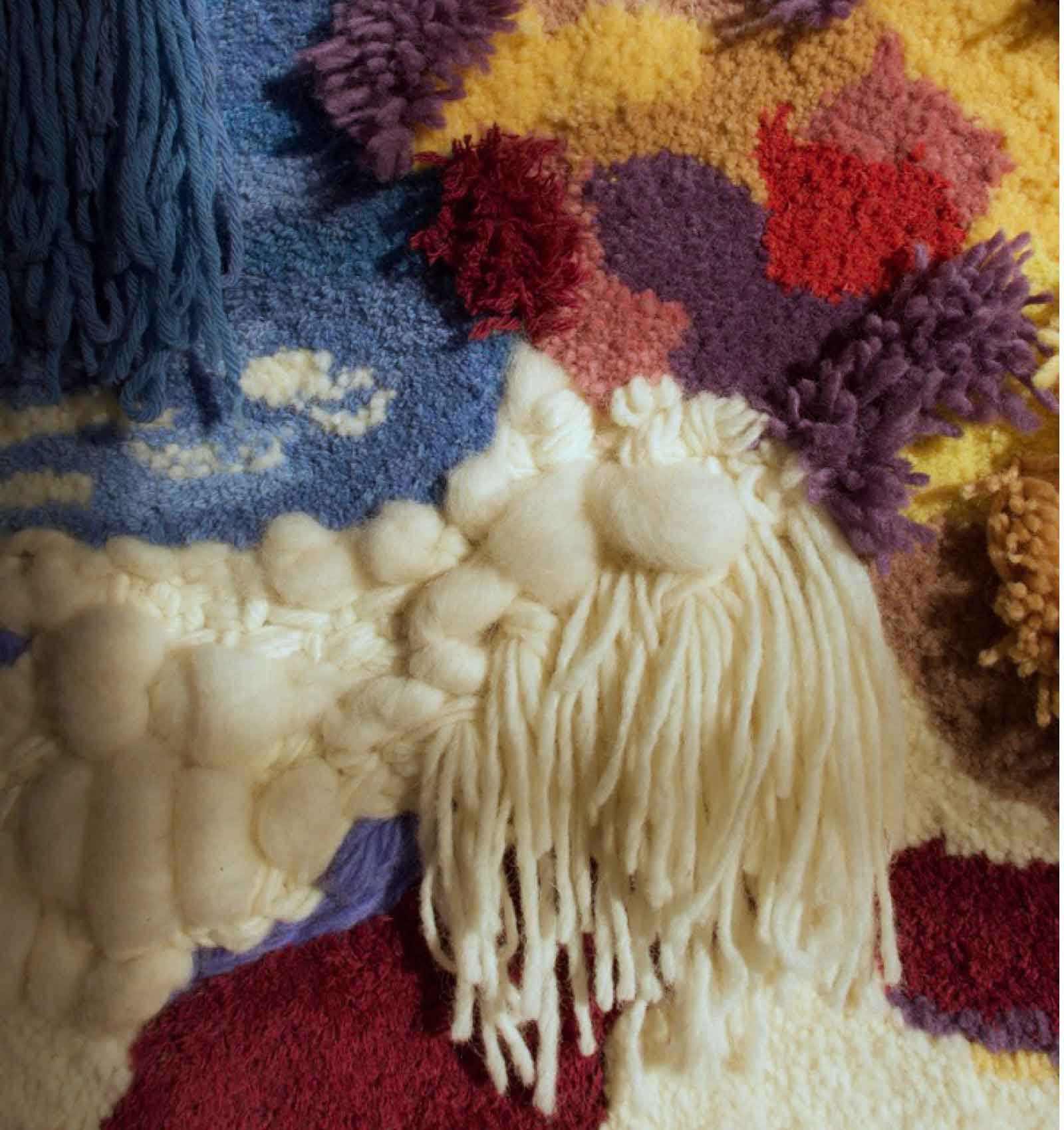 Close-up photo of a colorful woven rug with tassels and pom-poms, featuring patches of cream, red, purple, yellow, and blue colors.