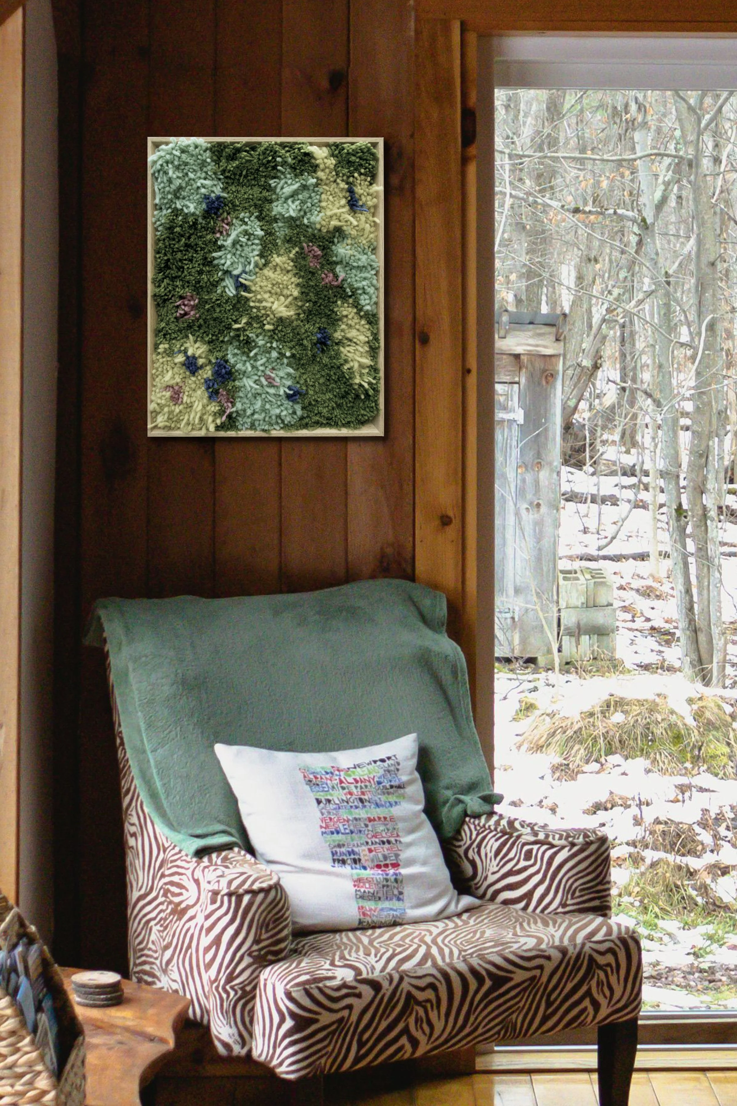 A cozy wooden room with a large window overlooking a snowy, leafless landscape. A zebra-print armchair with a green throw blanket and a white pillow with colorful writing is placed near the window. A framed textile art piece hangs on the wall above the chair.