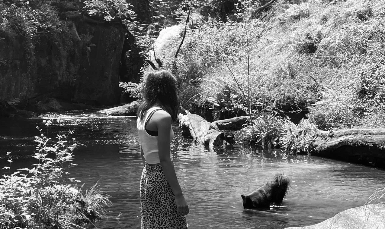 A woman standing by a river with a dog in the water, surrounded by trees and rocks in a natural setting.