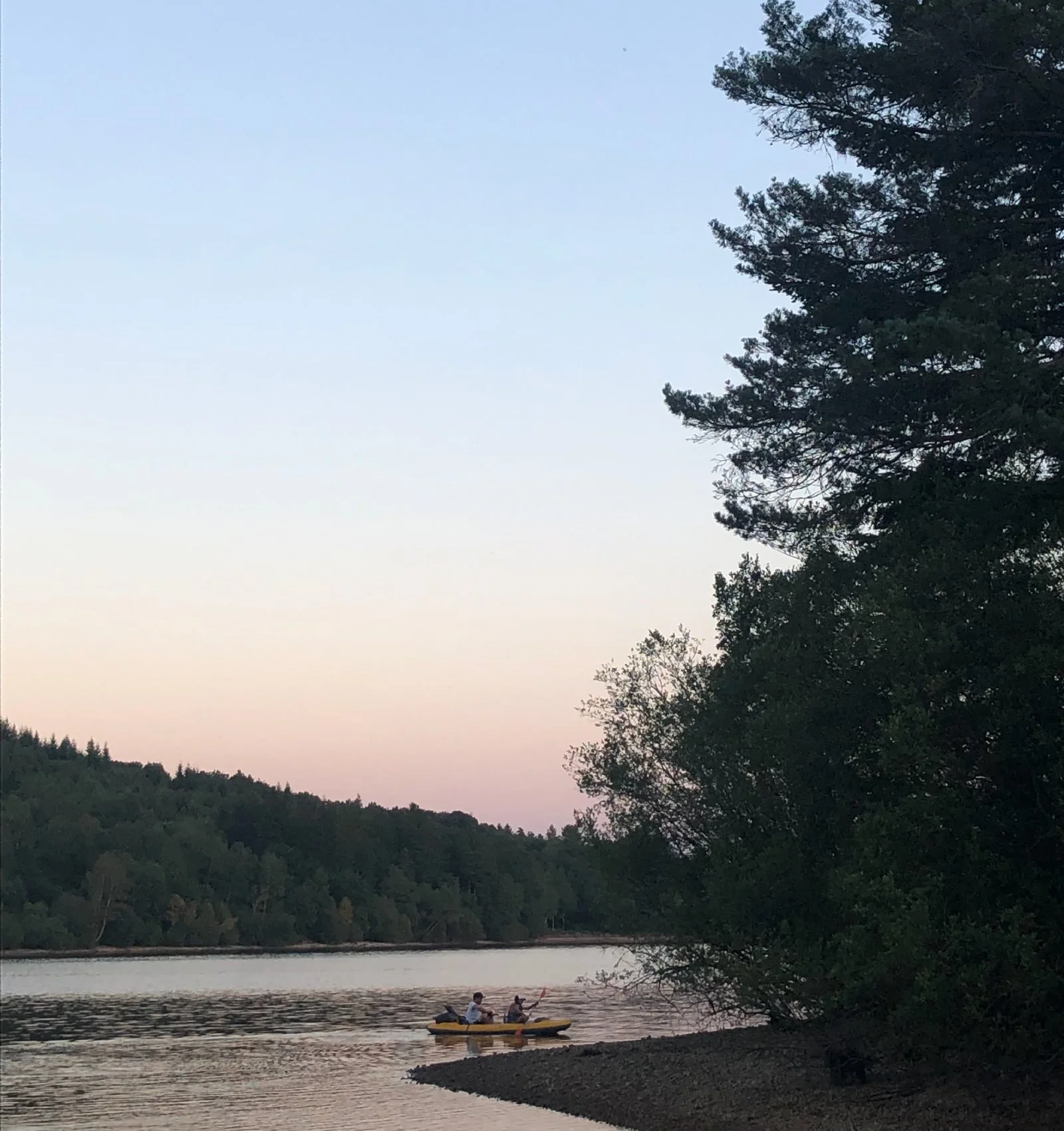People kayaking on a river near a forested shoreline at sunset, with trees on the right and hills in the distance.