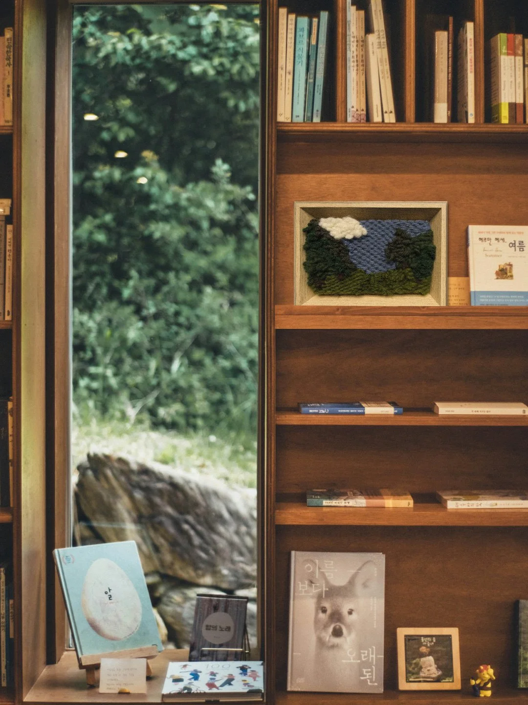 Bookshelf in a bookstore with books, a framed fiber art landscape, and children's books displayed near a window showing a green outdoor scene.