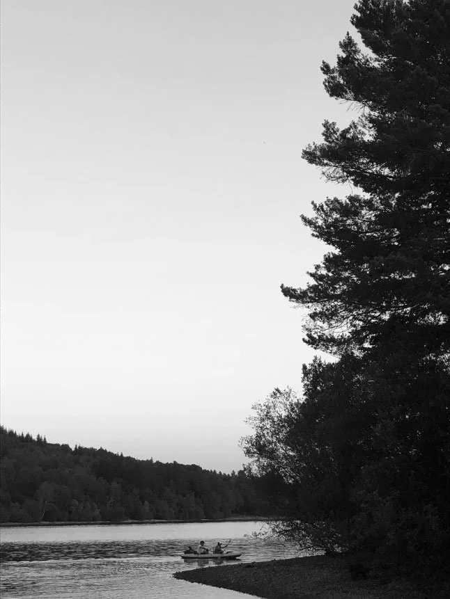 Black and white photo of a river with trees on the banks. In the distance, two people are in a small boat on the river.