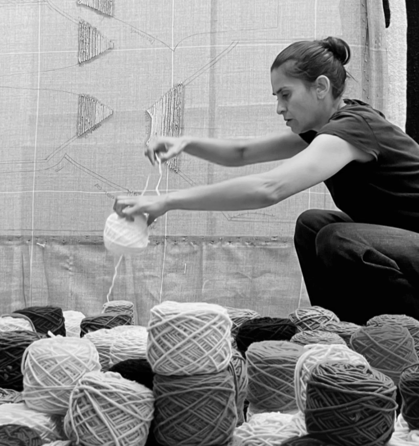 A woman crouches among numerous balls of yarn, handling one. Behind her is a wall with a large, geometric patterned quilt or textile art.