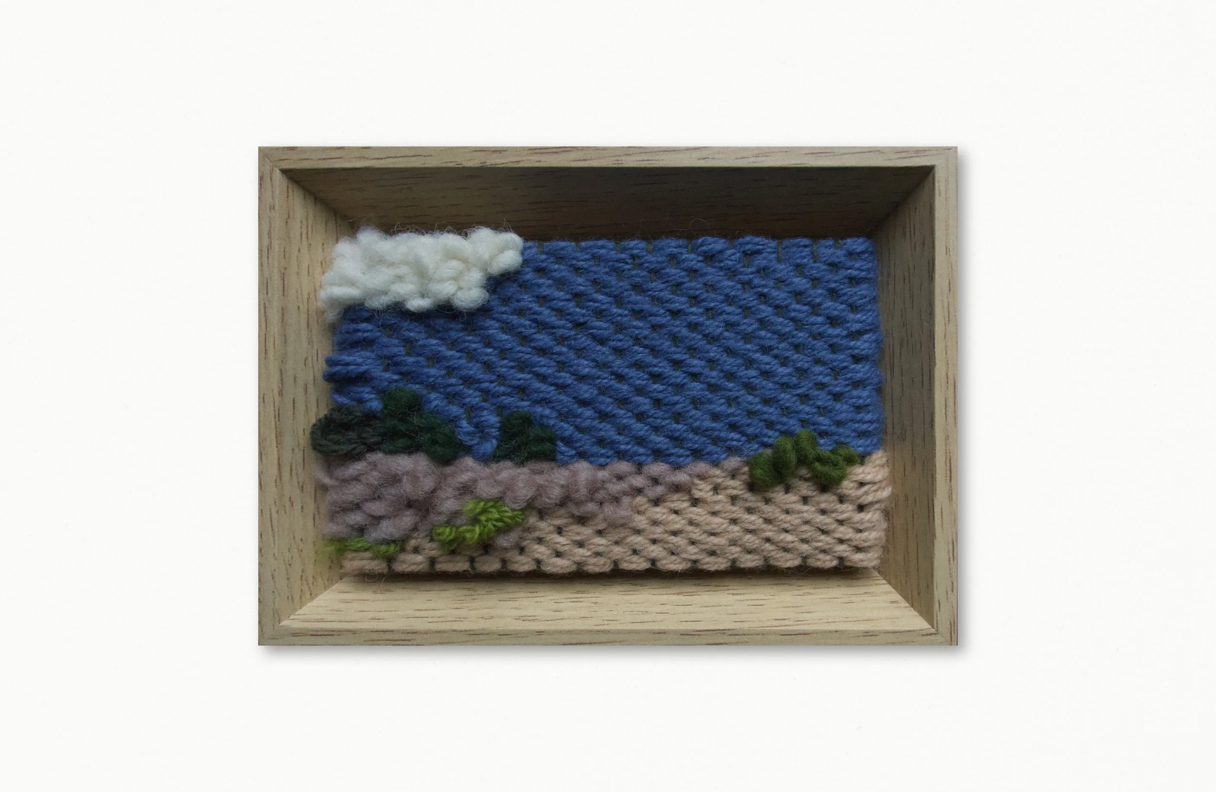 A framed piece of woven textile art with mountains, trees, and clouds, featuring various textures and colors, set in a light wooden box frame.